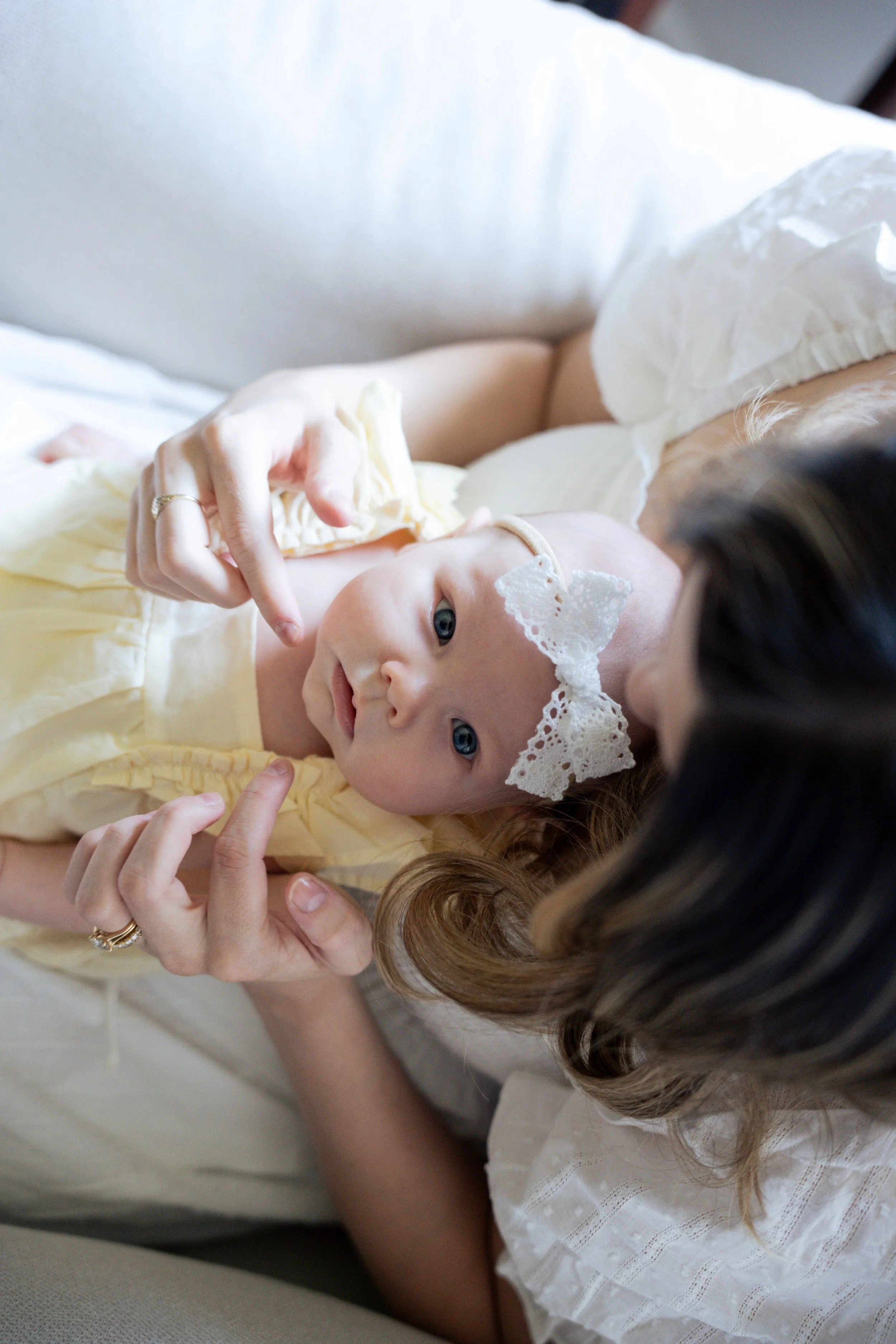 A woman with brown hair, wearing a white dress with lace, lying on a light-colored couch holding a baby girl with blue eyes and a light yellow dress. The baby has a white lace headband and is touching her face.