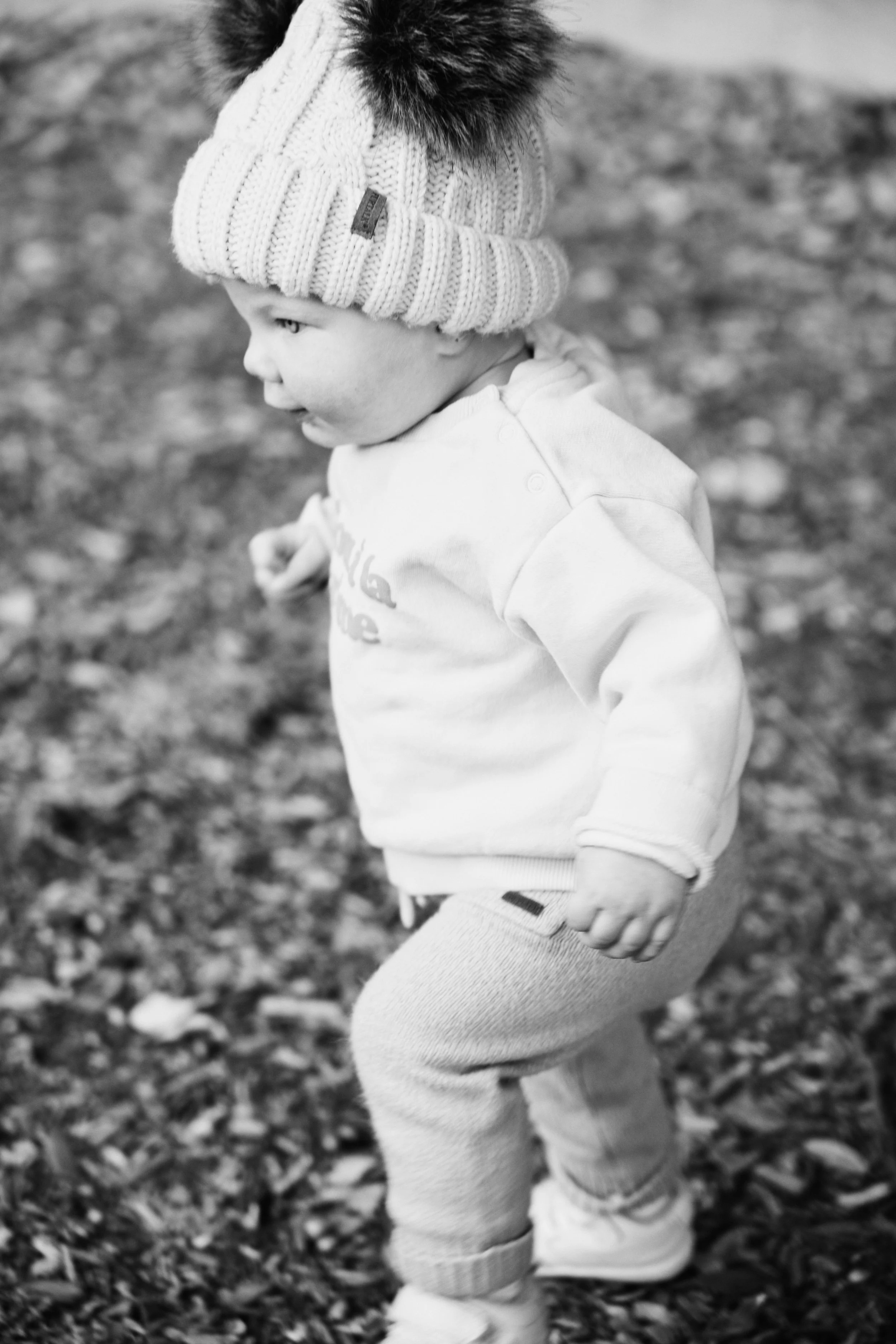 A young child wearing a knitted beanie with a furry pom-pom on top, a light-colored hoodie, and pants, standing outdoors on a leaf-covered ground, captured in profile in black and white.
