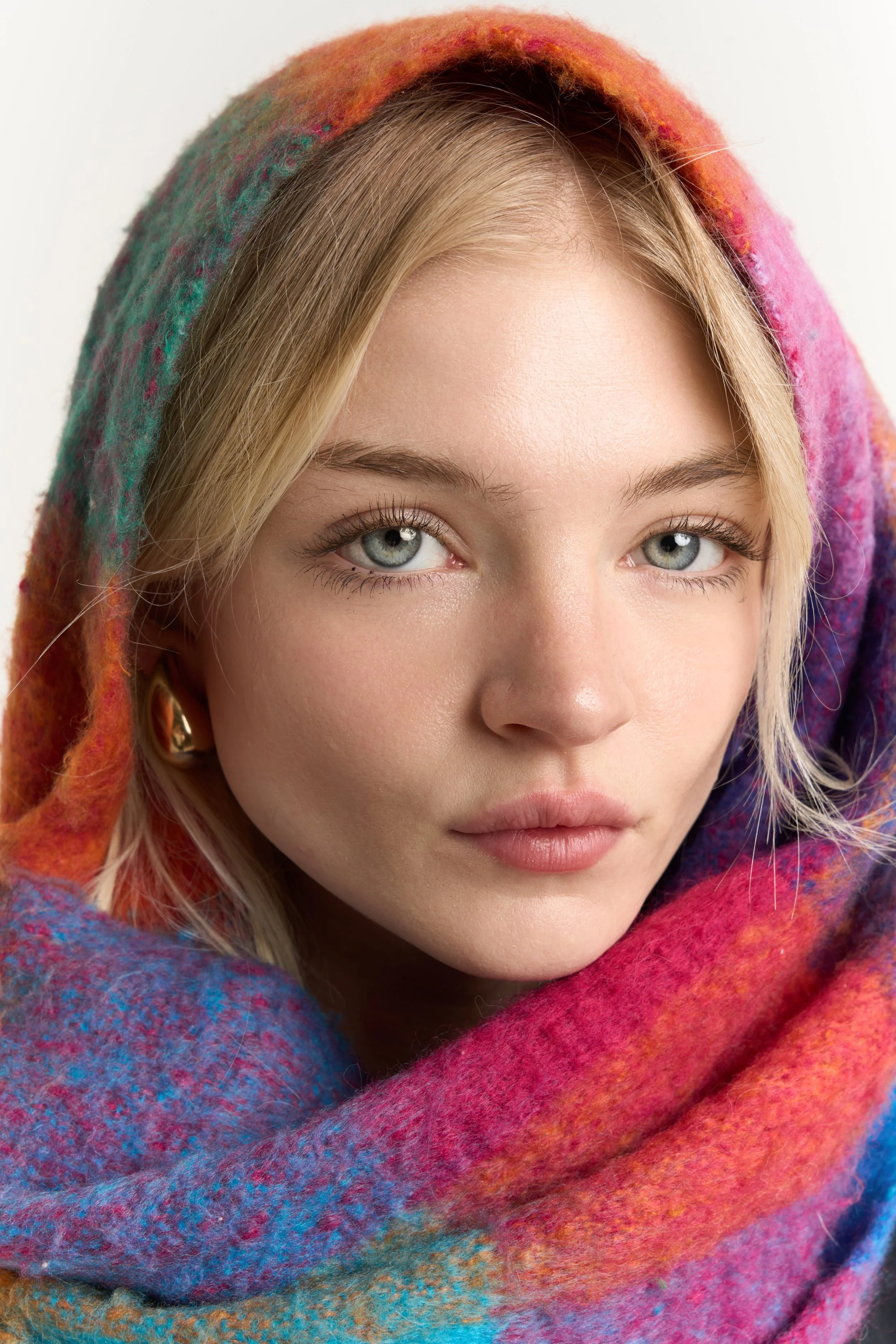 Close-up of a young woman's face with light blonde hair, fair skin, blue-gray eyes, and natural makeup, wrapped in a colorful, textured, rainbow-colored scarf or shawl.
