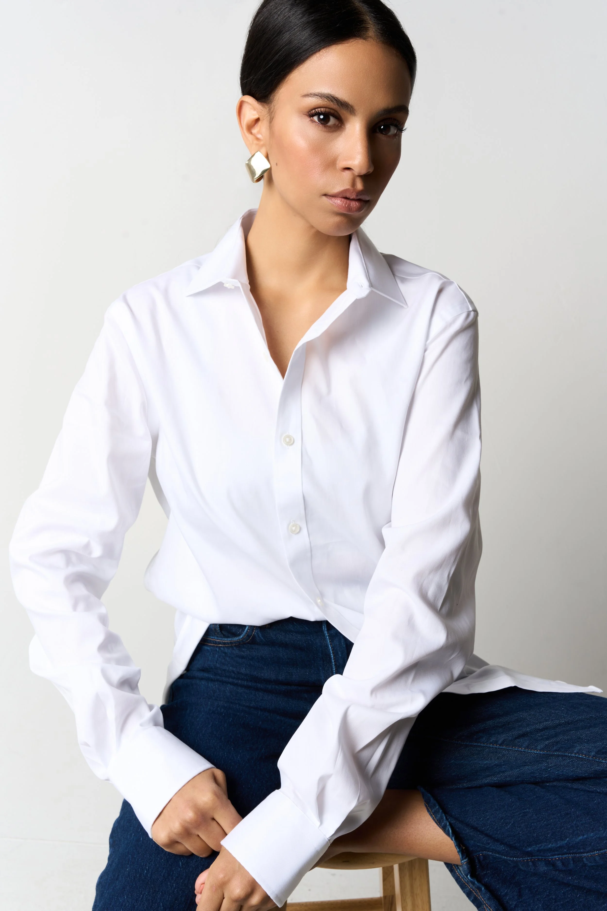 A woman with dark hair, wearing a white button-up shirt and dark blue jeans, sitting on a wooden stool against a plain white background.