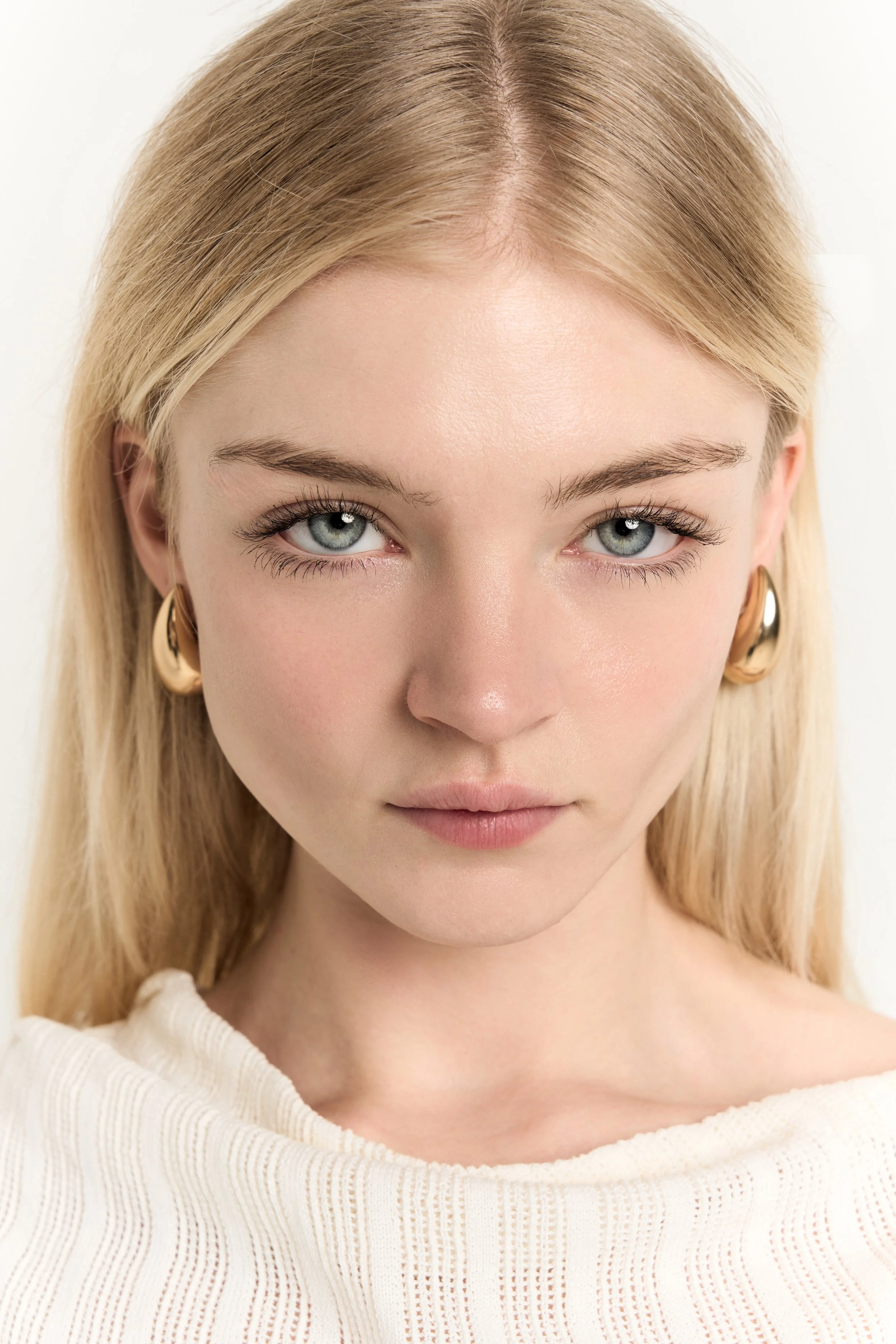 A close-up of a woman with blonde hair, blue eyes, and light skin, wearing a white sweater and gold earrings, looking directly at the camera.