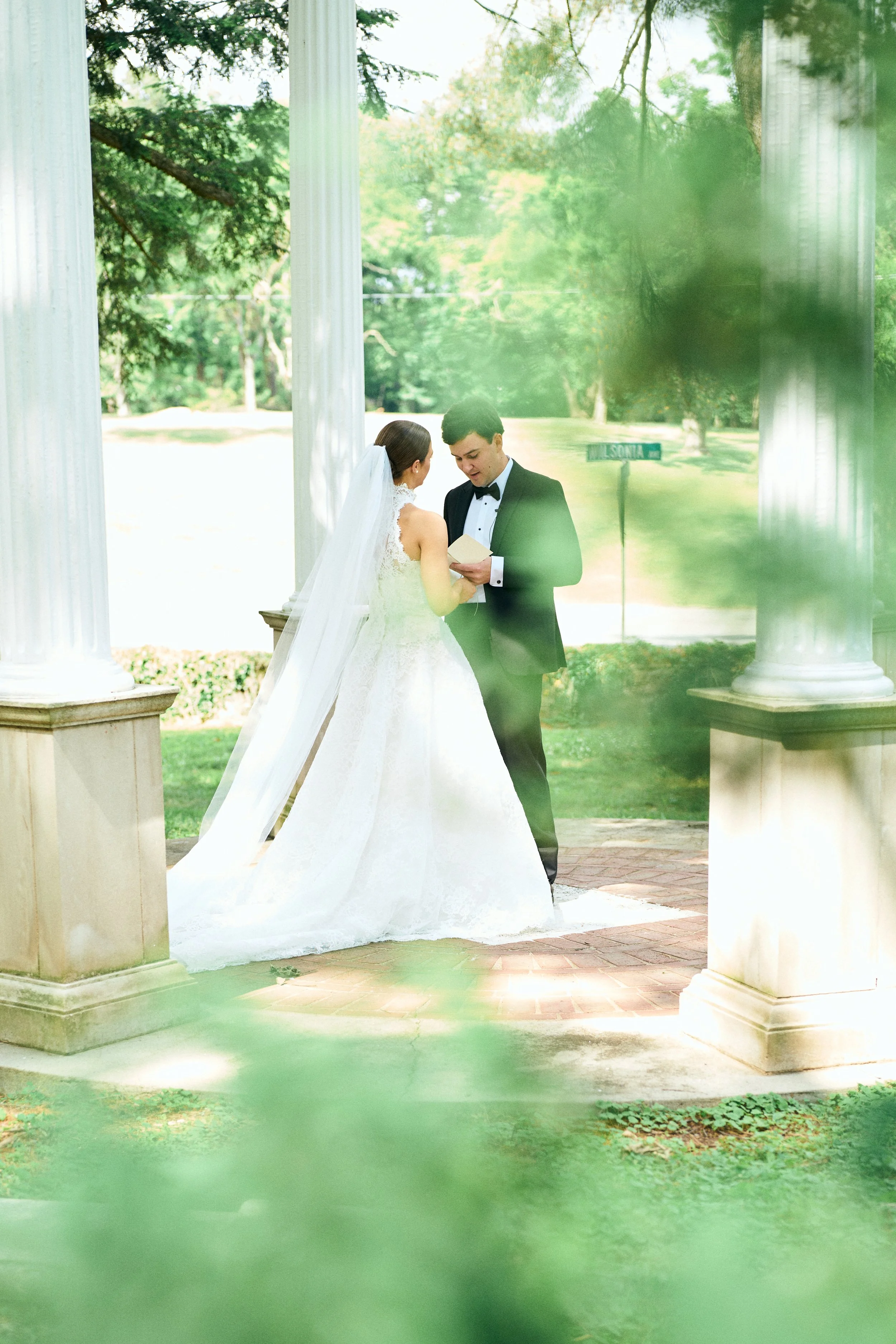 A bride and groom exchanging vows outdoors under a pavilion with white columns, green trees and a street sign in the background, seen through some greenery in the foreground.