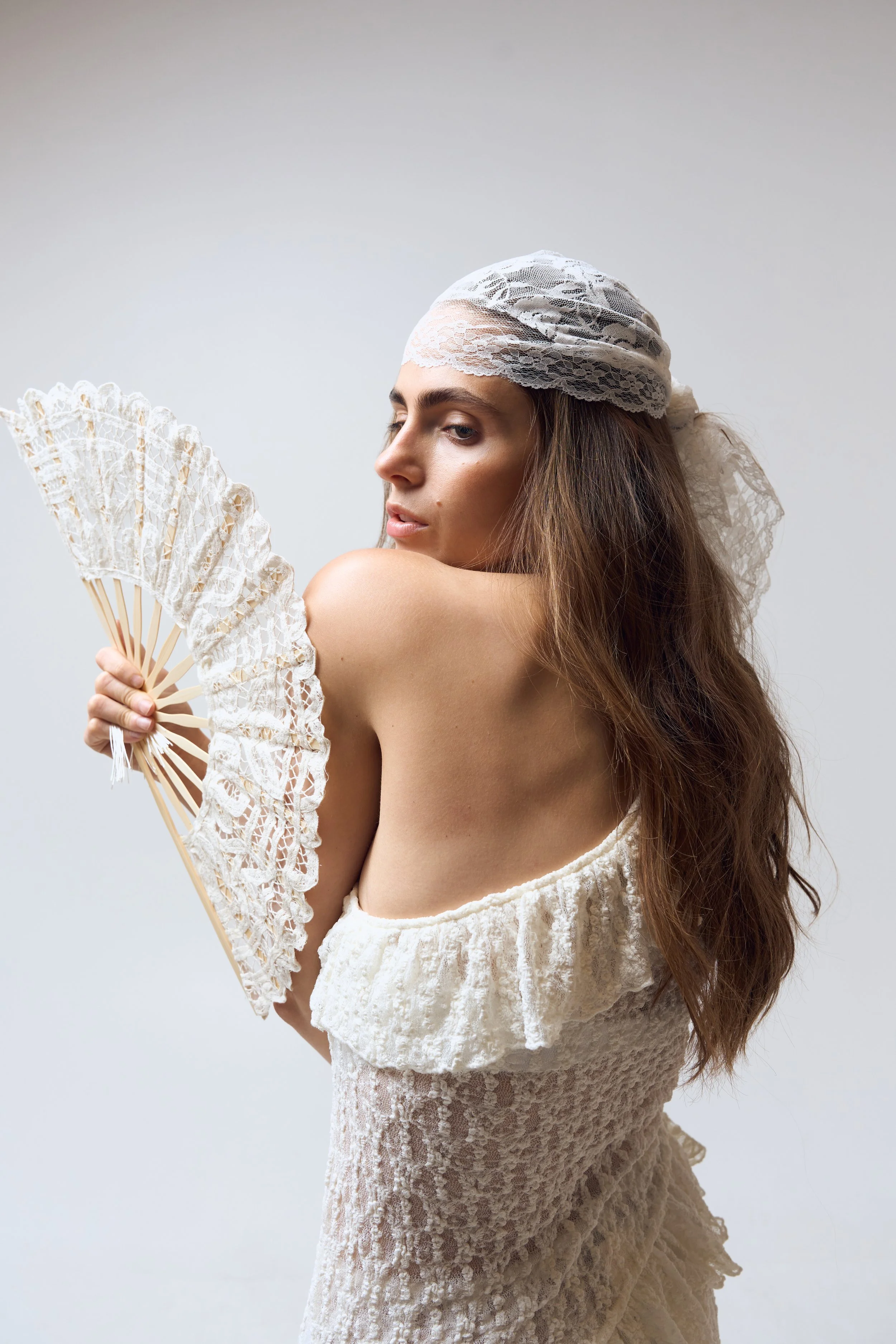 A woman with long brown hair wearing a white lace dress and a white lace head scarf, holding a white lace fan, posing against a plain backdrop.