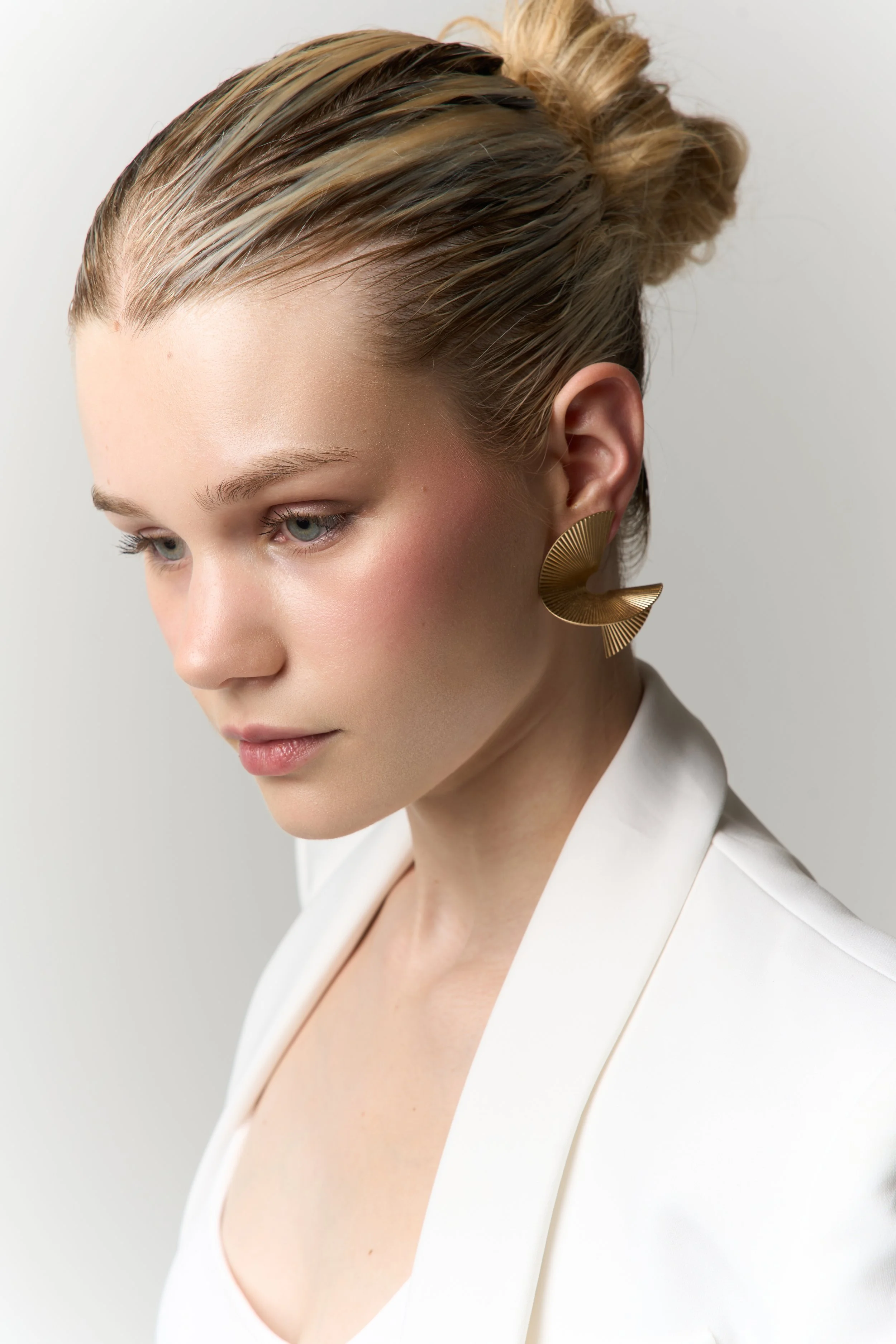A woman with blonde hair styled in a bun, wearing large gold earrings, a white blazer, and makeup with pink blush and natural lip color.