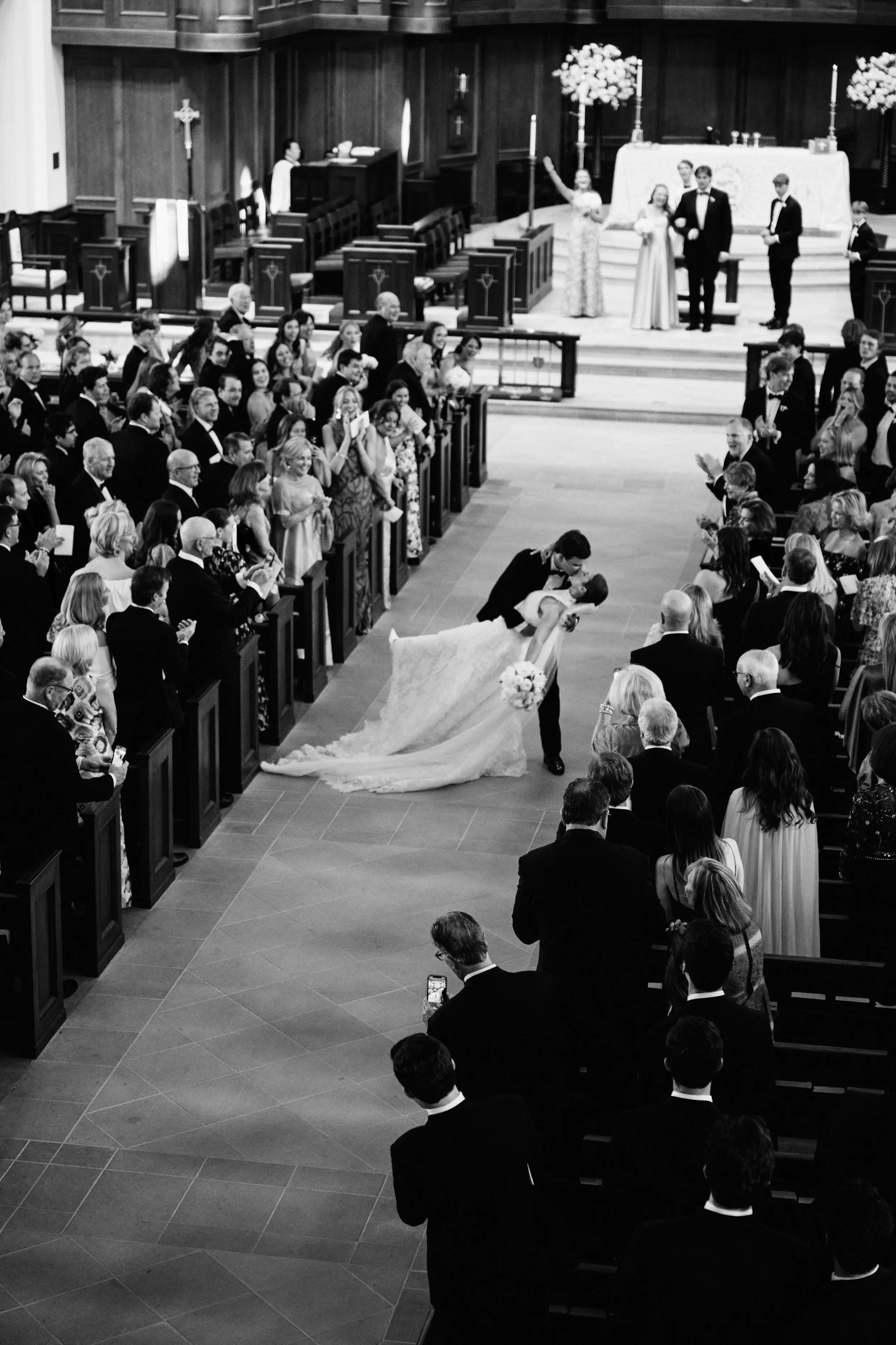 A wedding ceremony inside a church with guests dressed in formal attire, a bride and groom sharing a kiss while kneeling, and the officiant and wedding party standing at the altar.