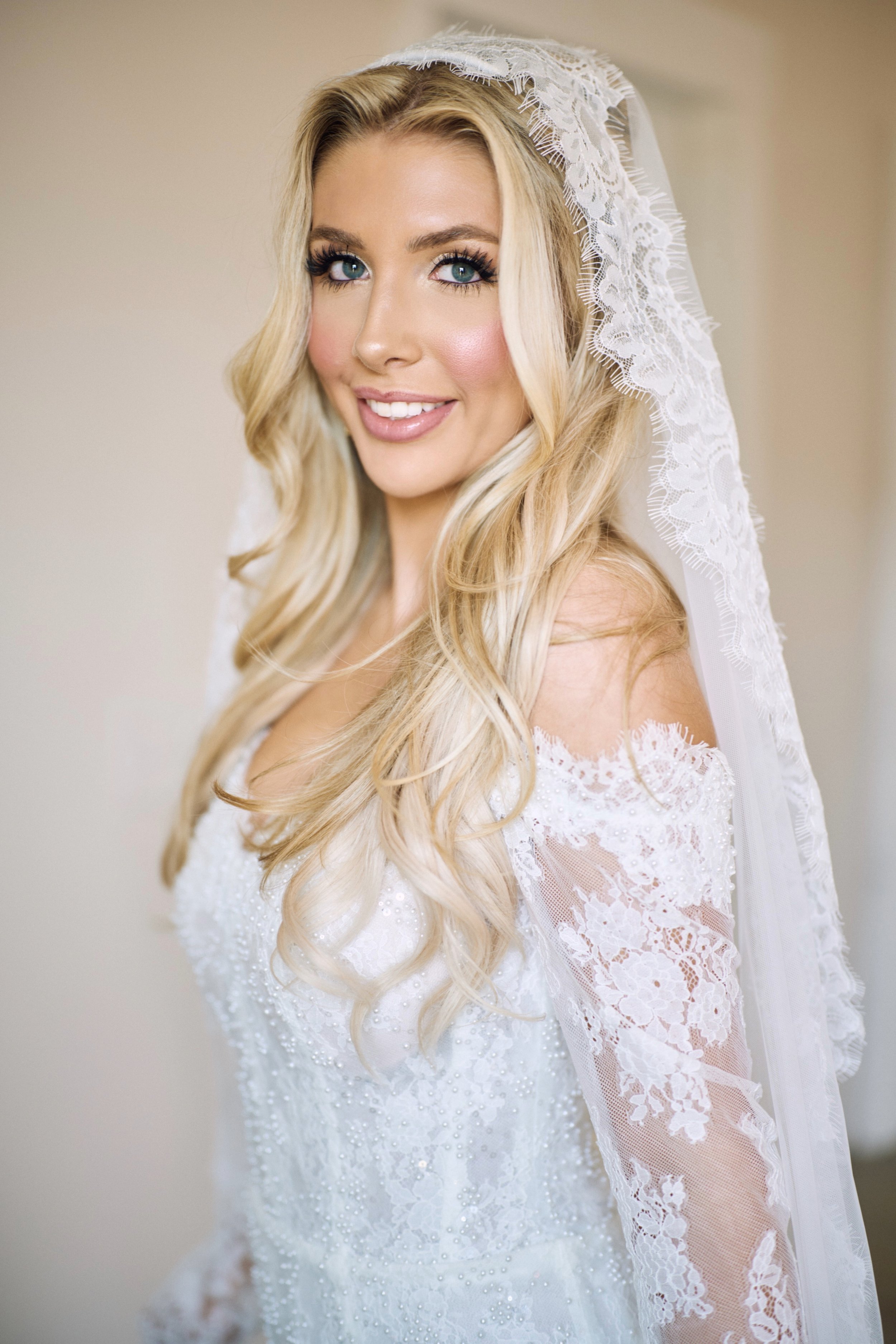 Close-up of a smiling woman with long blonde hair, blue eyes, wearing a white lace wedding dress and veil.