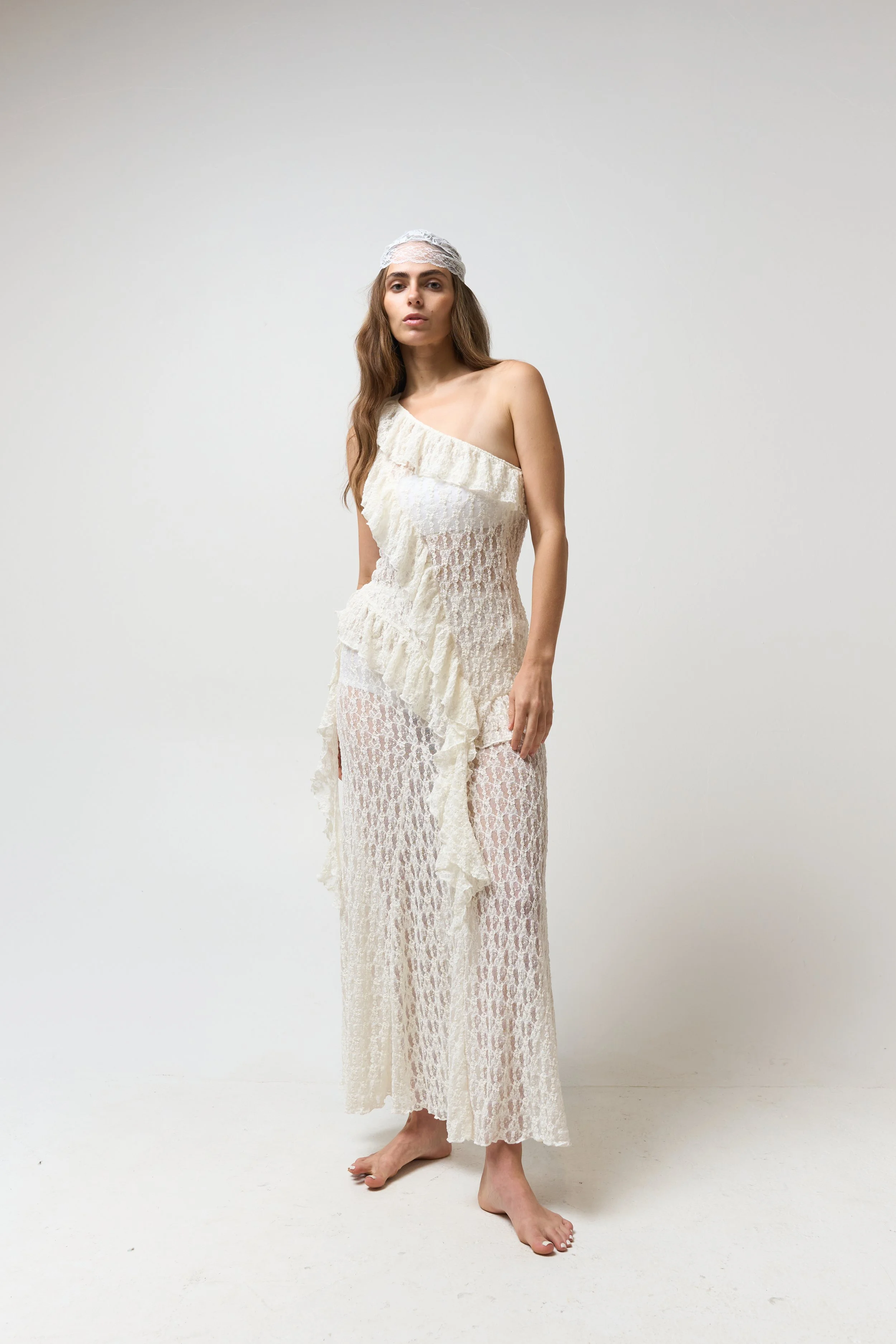 A woman stands barefoot against a plain white background, wearing a strapless, cream-colored lace dress with ruffles and a matching lace headscarf. She has long, wavy brown hair and a neutral expression.