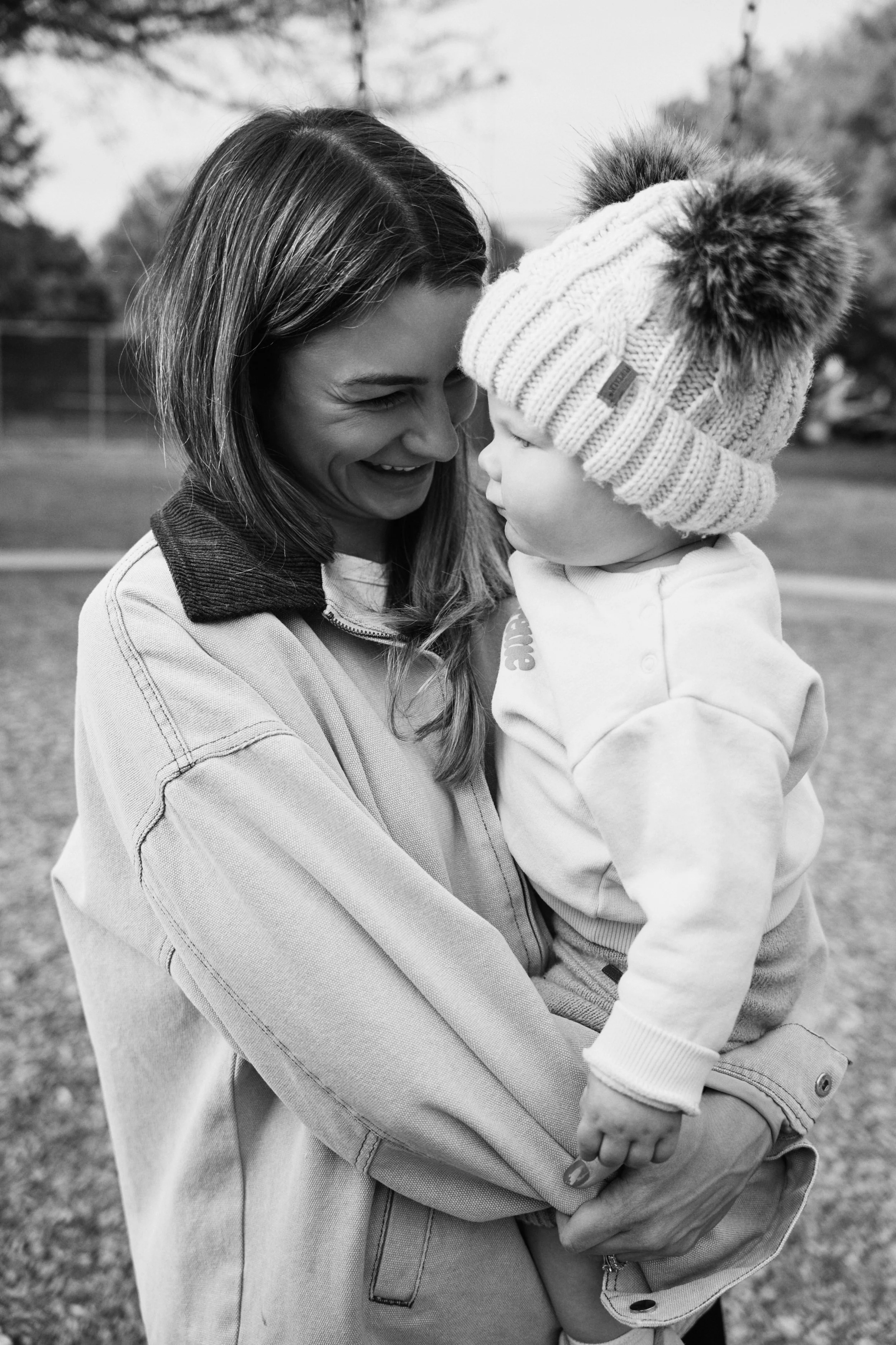 A woman and a young child are outdoors, close together, sharing a tender moment. The woman is smiling happily as she looks at the child, who is wearing a cozy knit hat with a pom-pom and a light-colored jacket. The woman is wearing a light jacket wit
