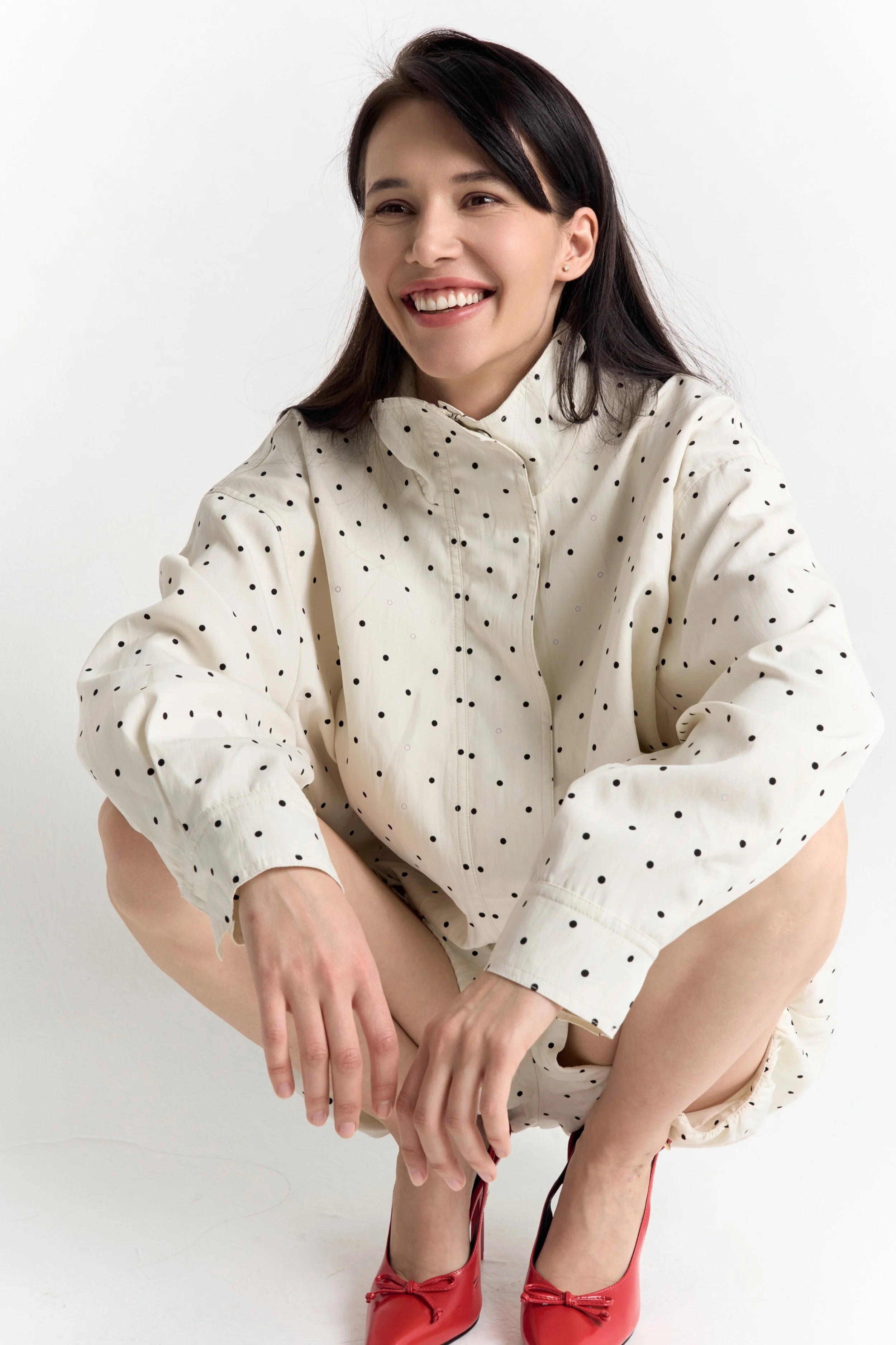 A woman with dark hair and light skin, smiling, wearing a white polka dot outfit and red heels, posing against a plain white background.