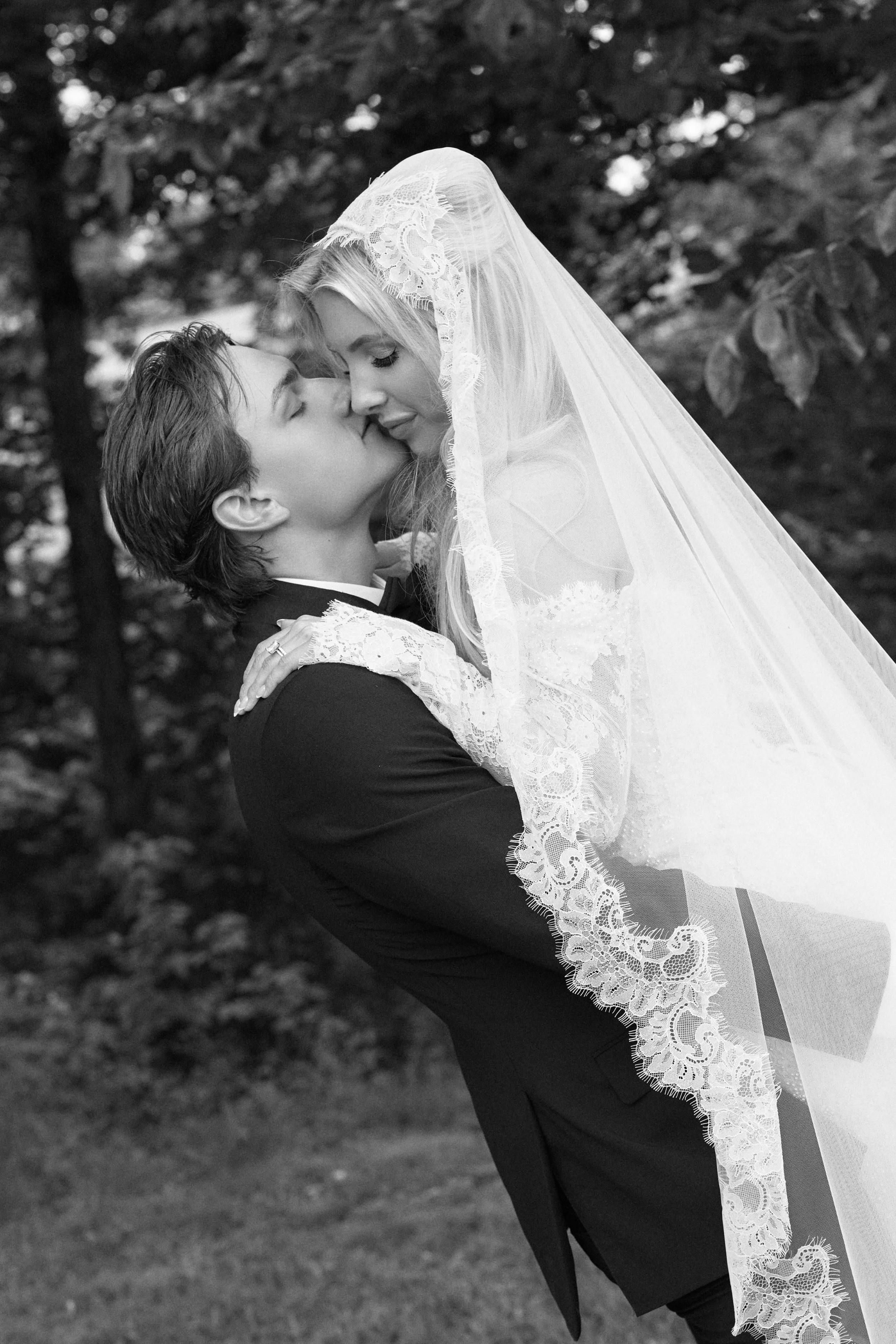 A bride and groom embrace outdoors, with the groom lifting the bride. The bride wears a lace wedding veil and dress, and the groom wears a dark suit. They are surrounded by trees.
