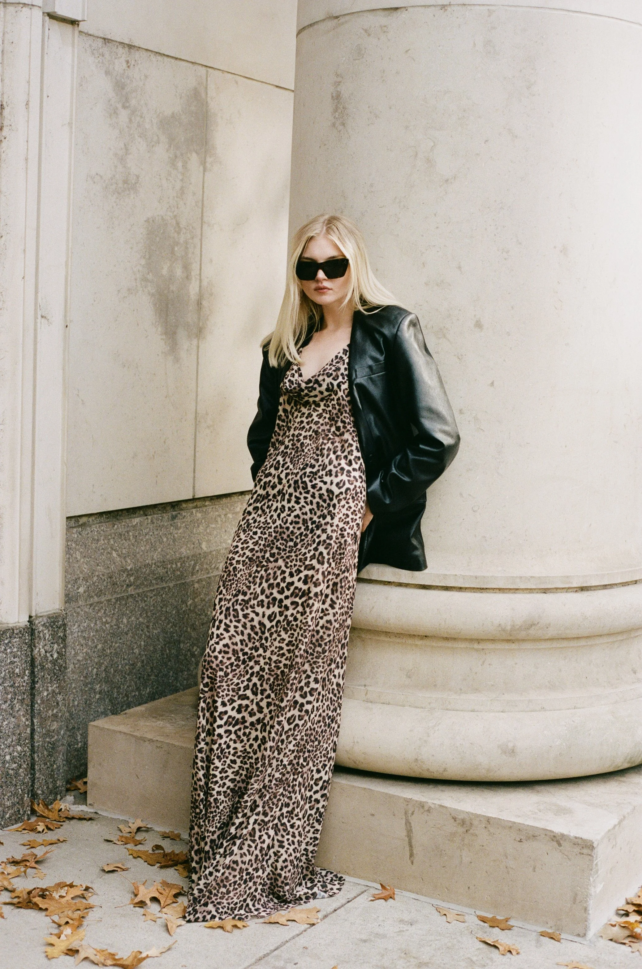 A woman with blonde hair wearing black sunglasses, a leopard print dress, and a black leather jacket stands in front of a concrete column on a sidewalk with fallen leaves.