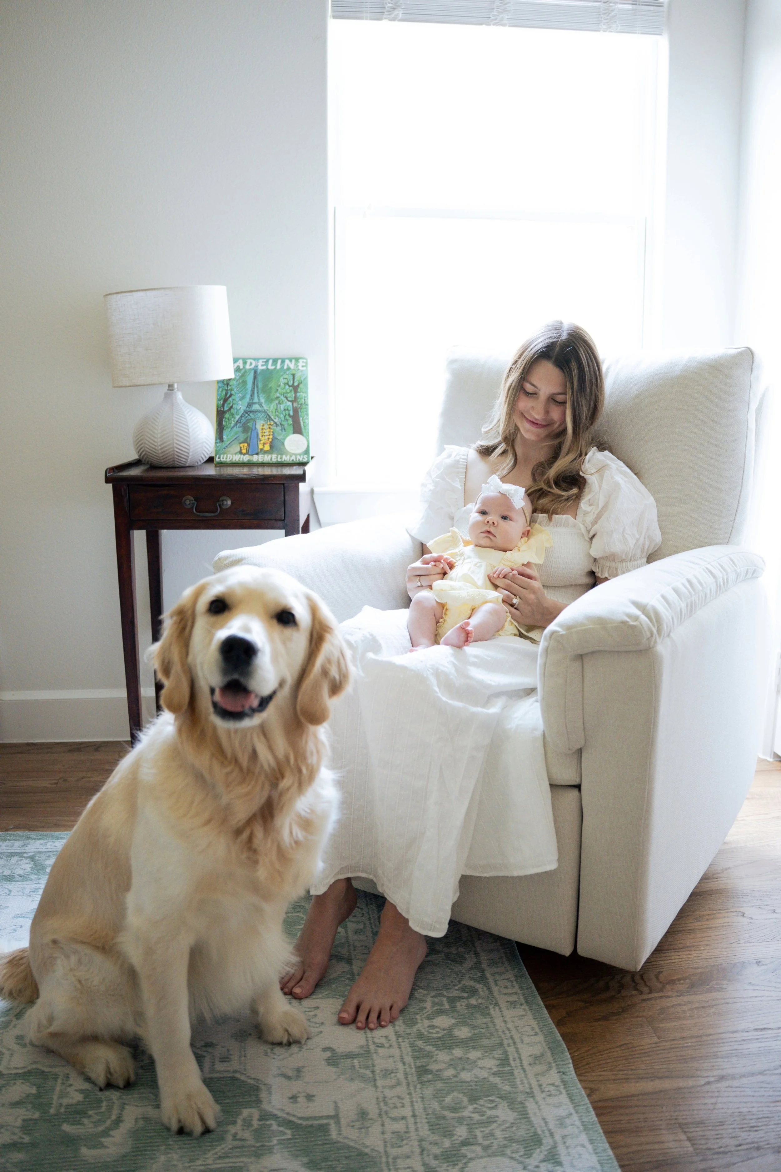 A woman sitting on a cream-colored armchair holding a baby dressed in yellow, with a dog sitting on a rug in front of her. A window with blinds and a nightstand with a lamp and a colorful book are in the background.