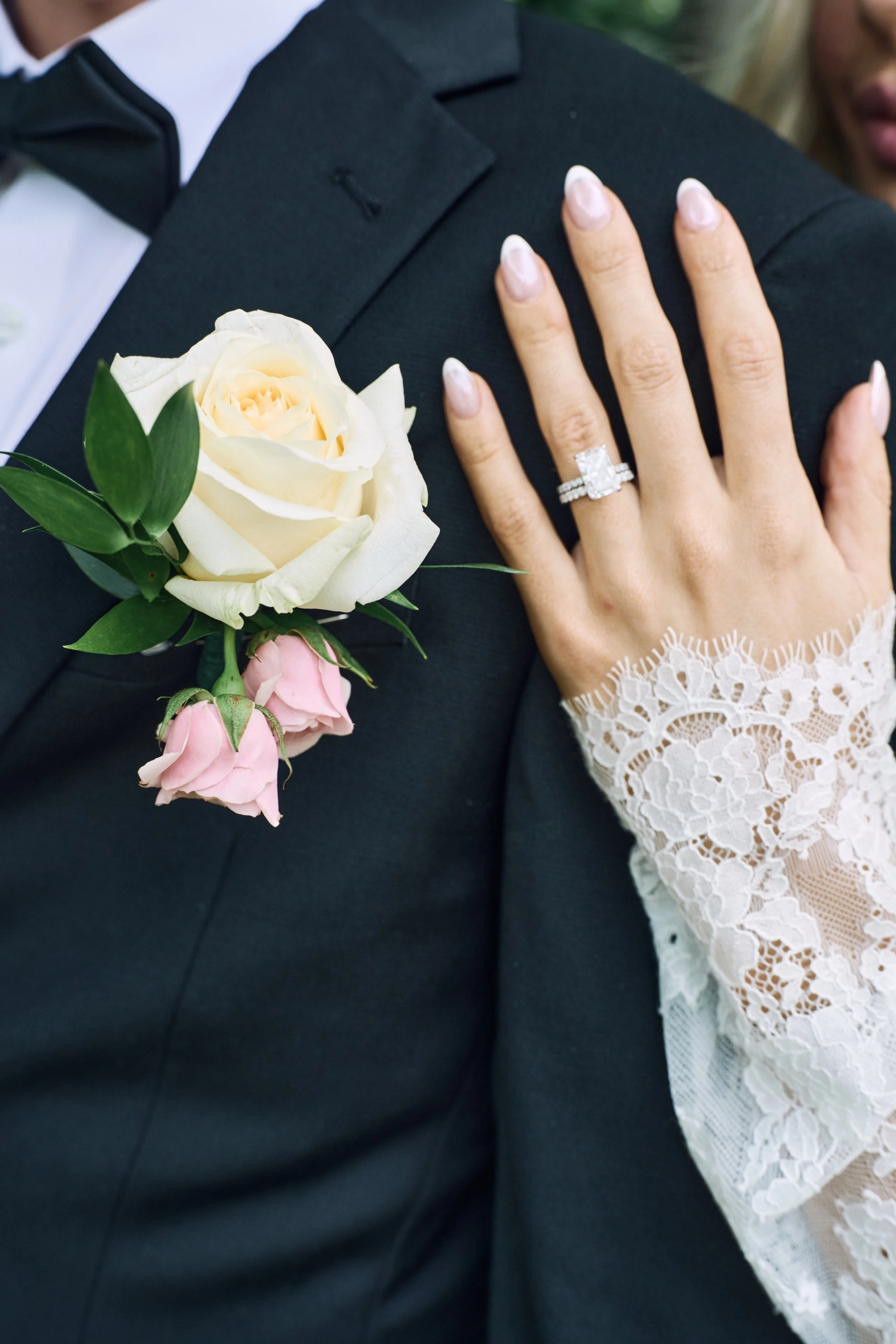 A close-up of a bride and groom at their wedding. The groom is wearing a black tuxedo with a white shirt and black bow tie. The bride's left hand, with a diamond engagement ring, is resting on the groom's shoulder. The bride is wearing a white lace w
