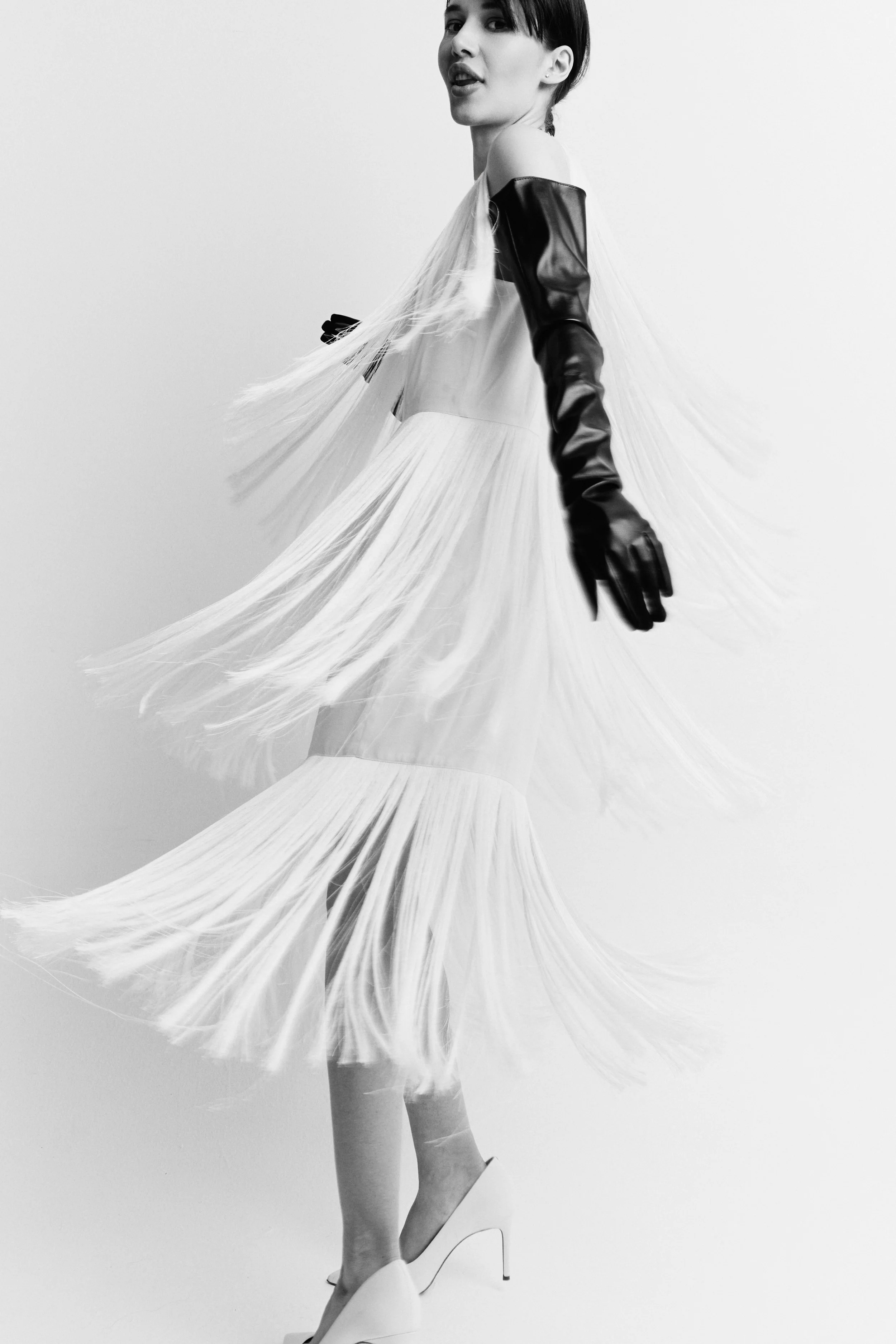 Black and white photo of a woman wearing a light-colored tulle dress with a fluffy skirt, a satin top, and black long gloves, standing on high heels against a plain background.