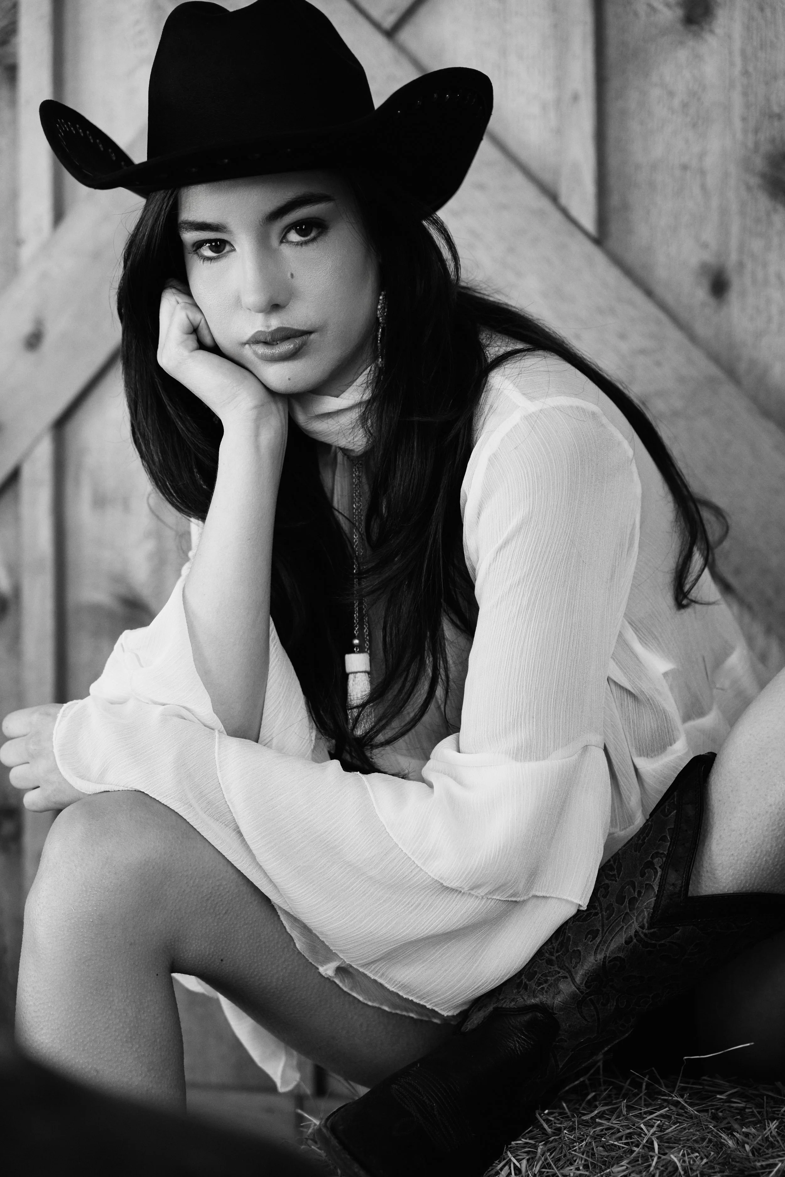 A woman wearing a cowboy hat and a long-sleeve shirt sitting on straw or hay, with her chin resting on her hand, looking directly at the camera in a black and white photo.