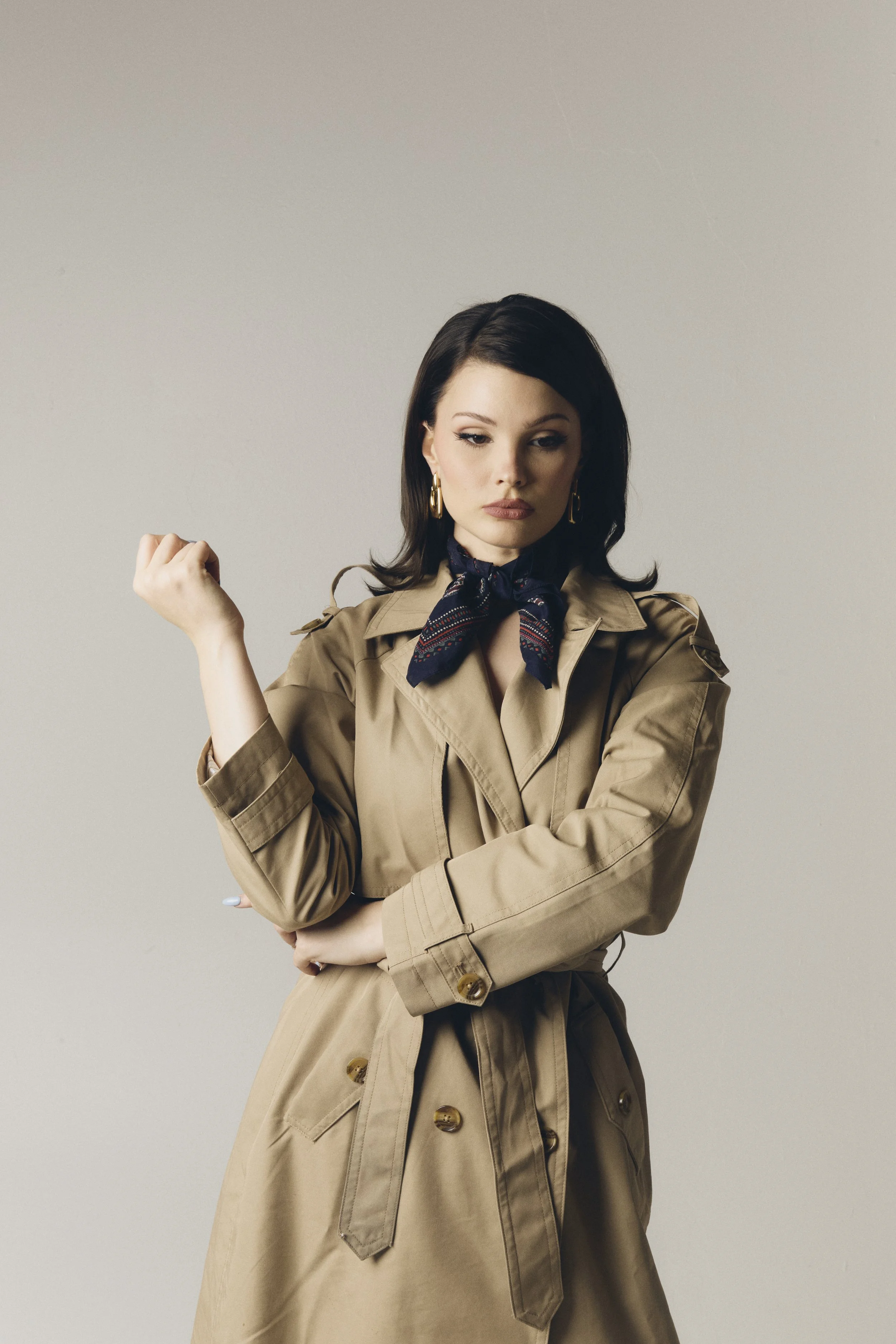 A woman with dark hair wearing a beige trench coat, a navy blue patterned scarf, and gold hoop earrings, standing with her arms crossed and looking pensively to the side against a plain light gray background.