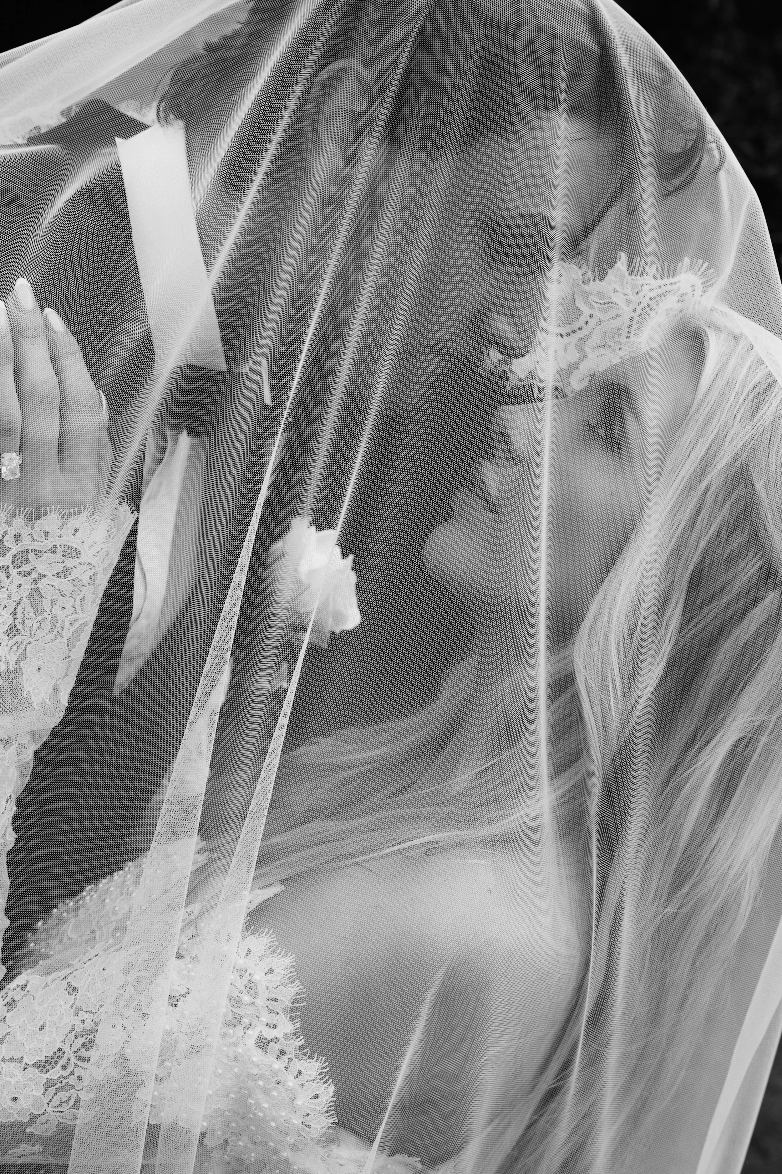 A bride with a veil over her face, wearing lace wedding dress, holding a bouquet, smiling.