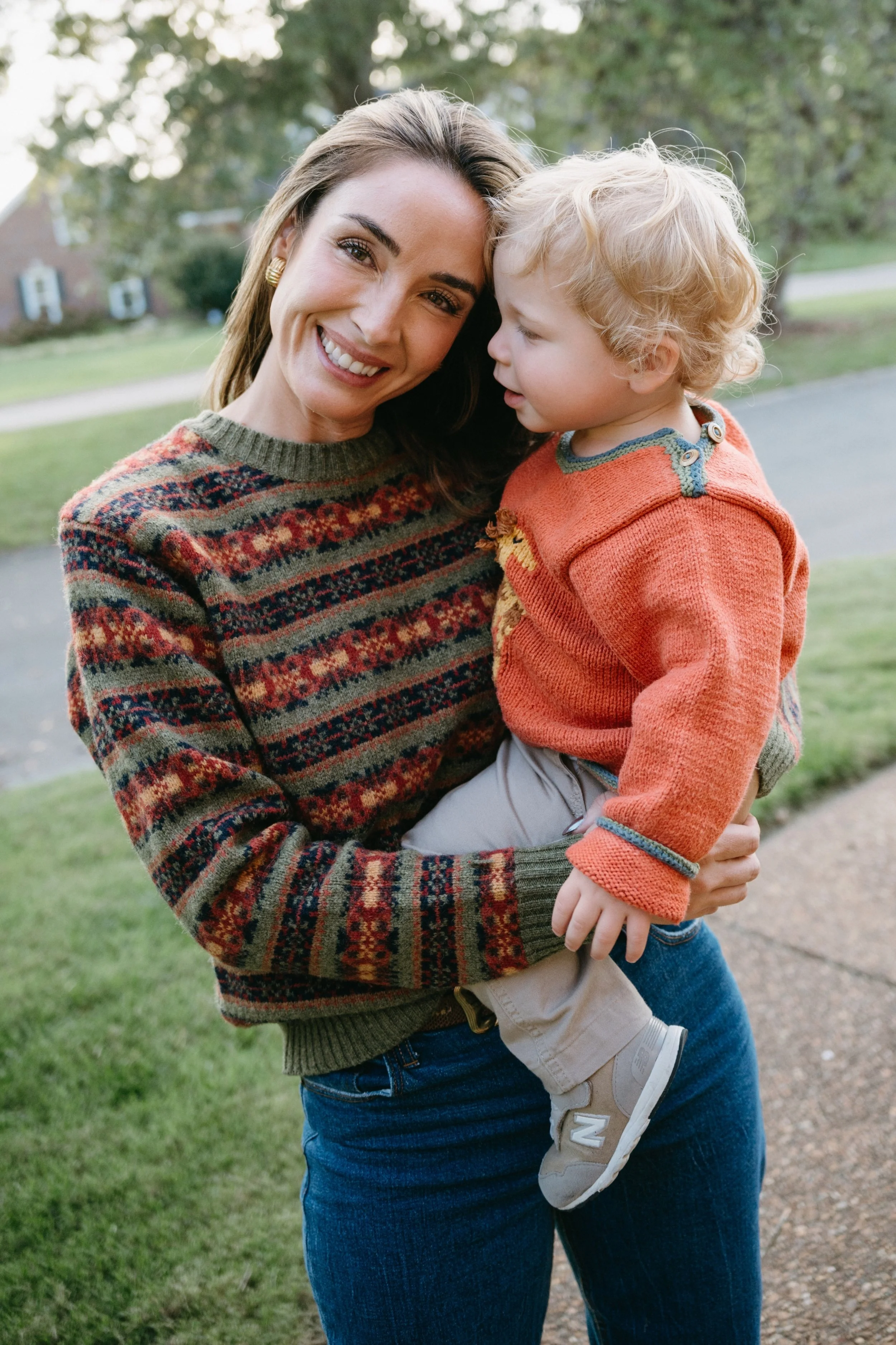 A woman smiling and holding a young boy outdoors in a park with green grass and trees in the background.