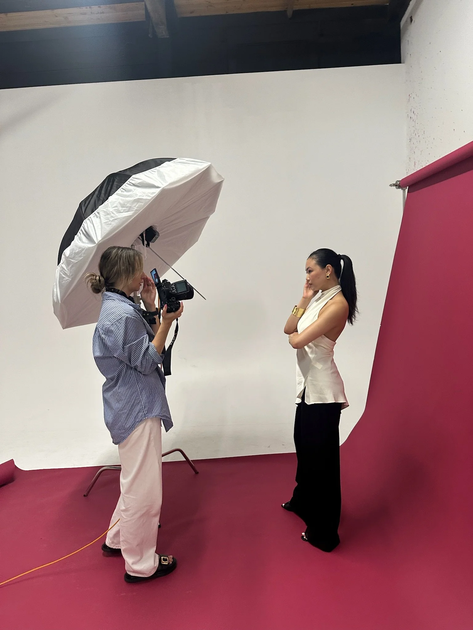 A photography studio with a photographer taking a picture of a female model. The photographer uses a large umbrella light to illuminate the model, who is standing against a plain white background with a pink backdrop nearby.