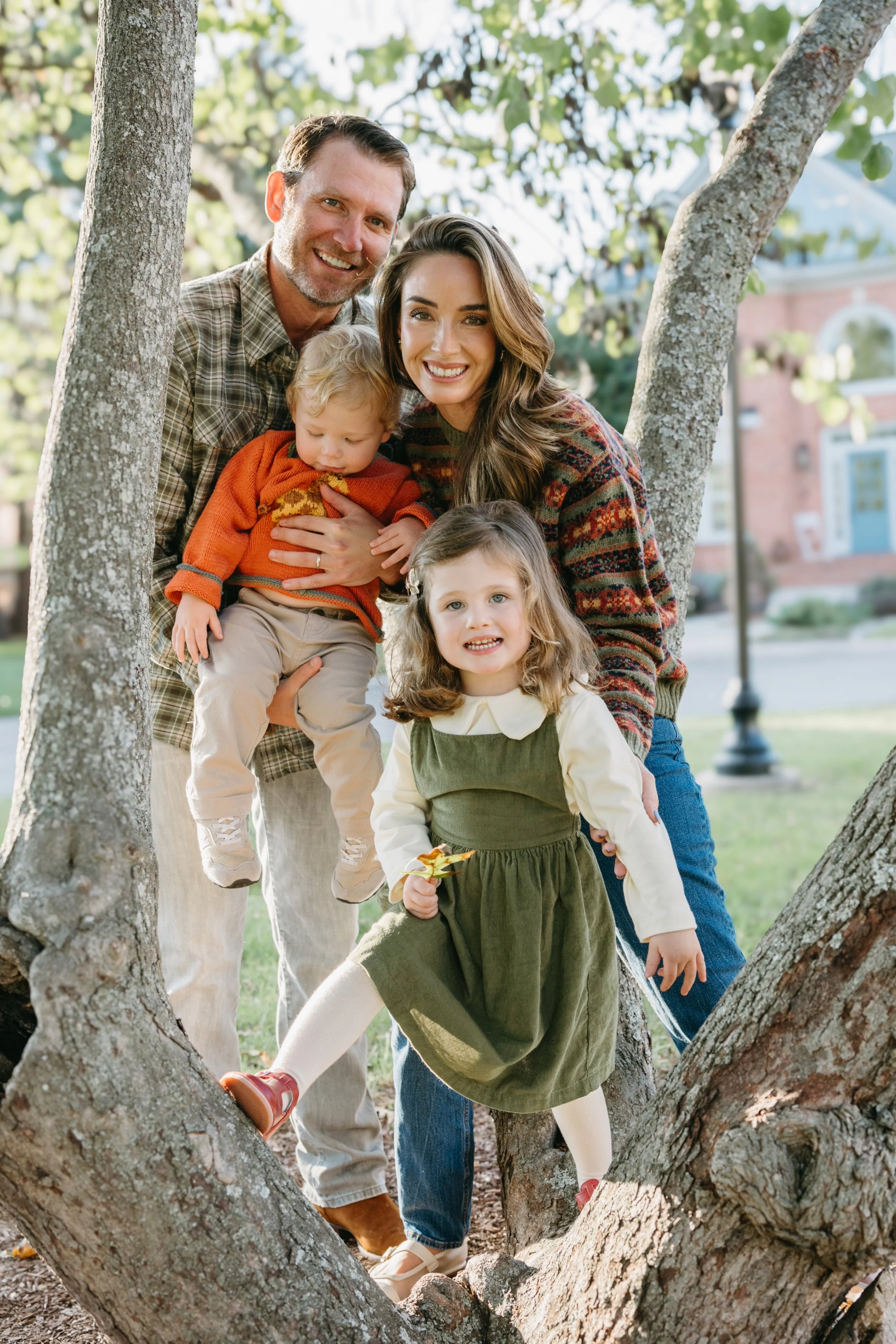 A smiling family of four, including a father, mother, and two young daughters, enjoying time outdoors by a large tree in a park.