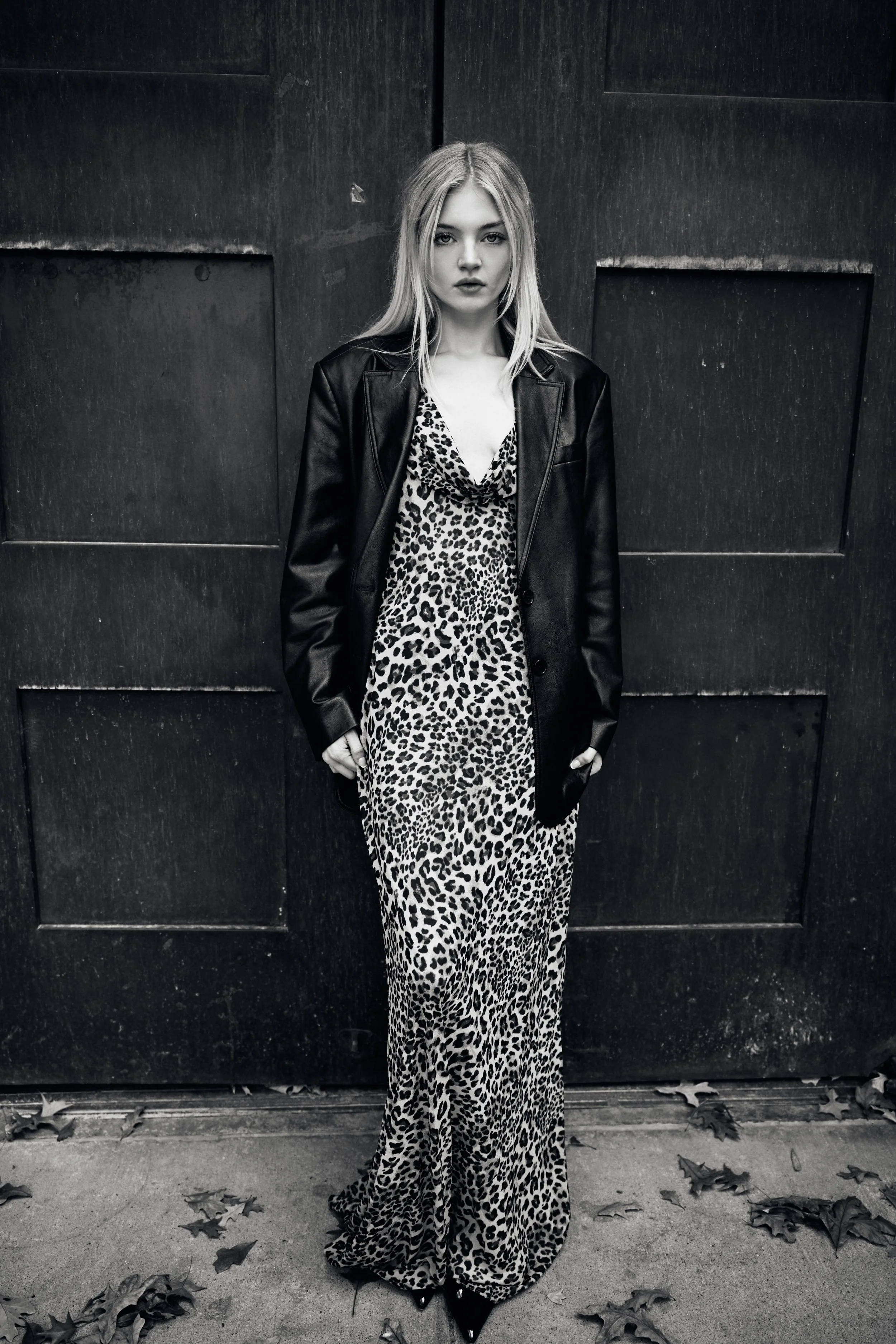 A woman standing against a dark wooden door, wearing a leopard print dress with a low neckline, a black leather jacket, and black shoes. She has long blonde hair and a serious expression.