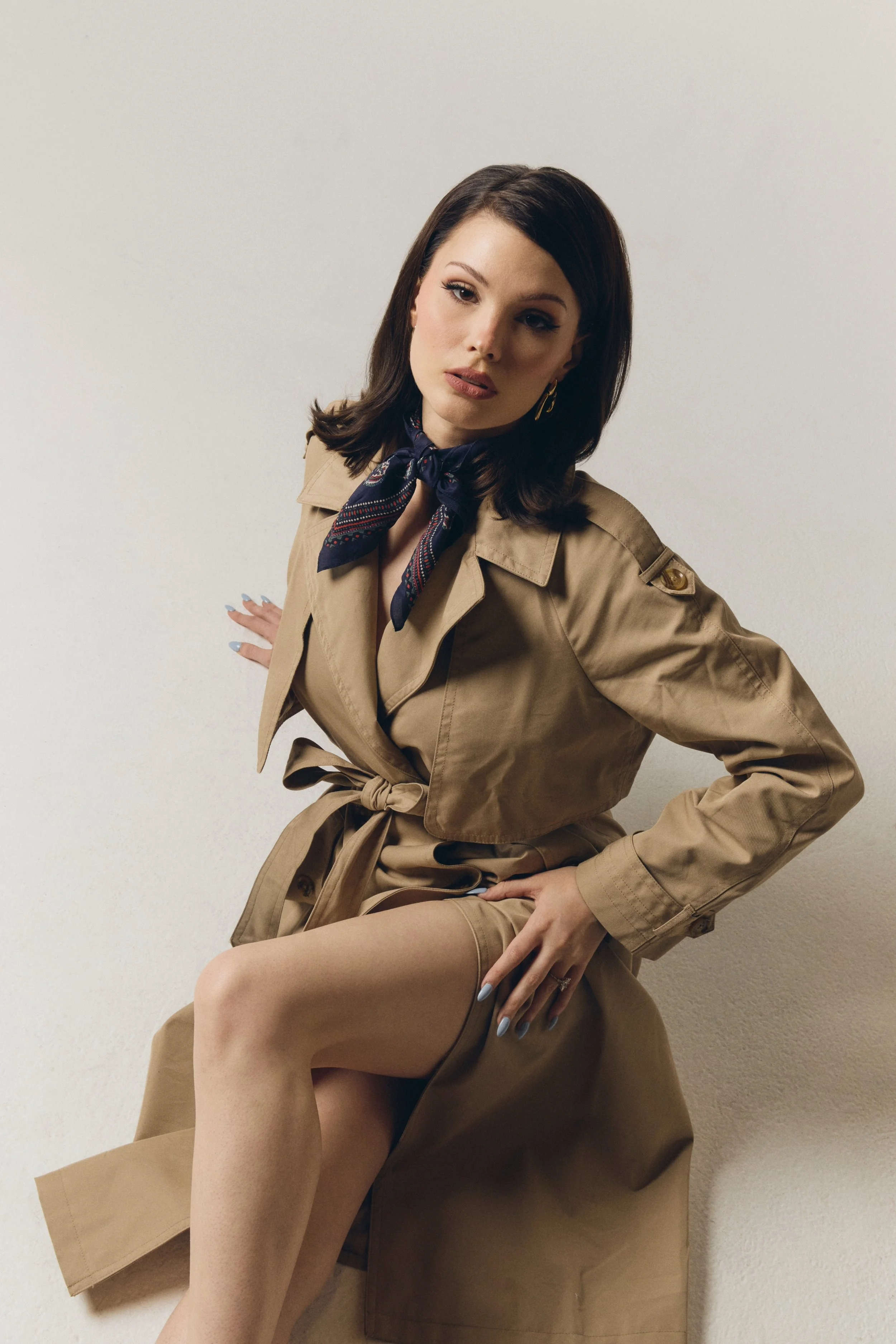 A woman with dark brown hair in a beige trench coat, blue scarf, and beige stockings, sitting against a plain off-white wall, with one hand on her hip and the other resting near her knee.