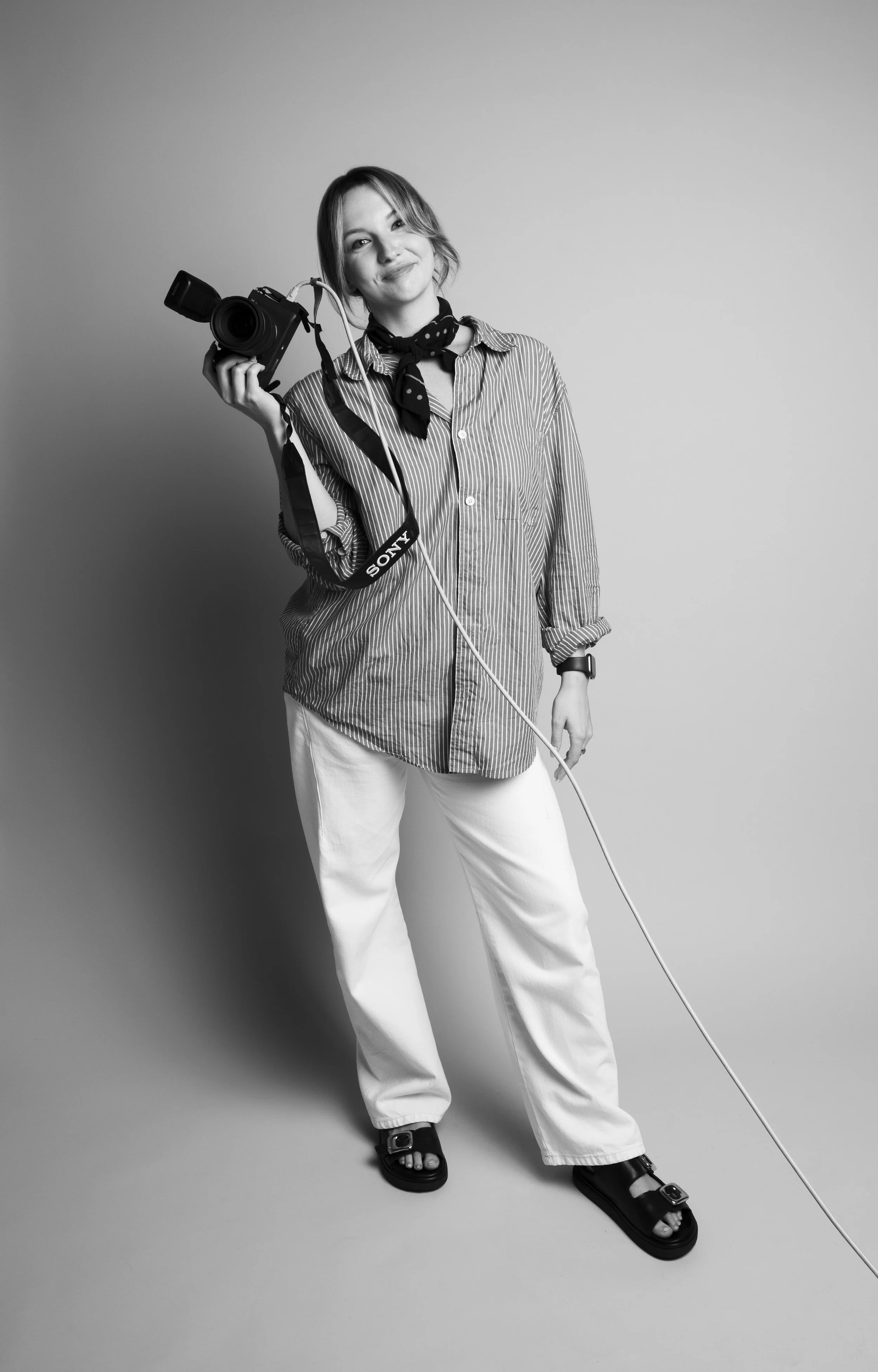 Black and white photo of a woman with short hair, wearing a striped shirt, white pants, and sandals, holding a camera with a charging cable in front of a blank background.