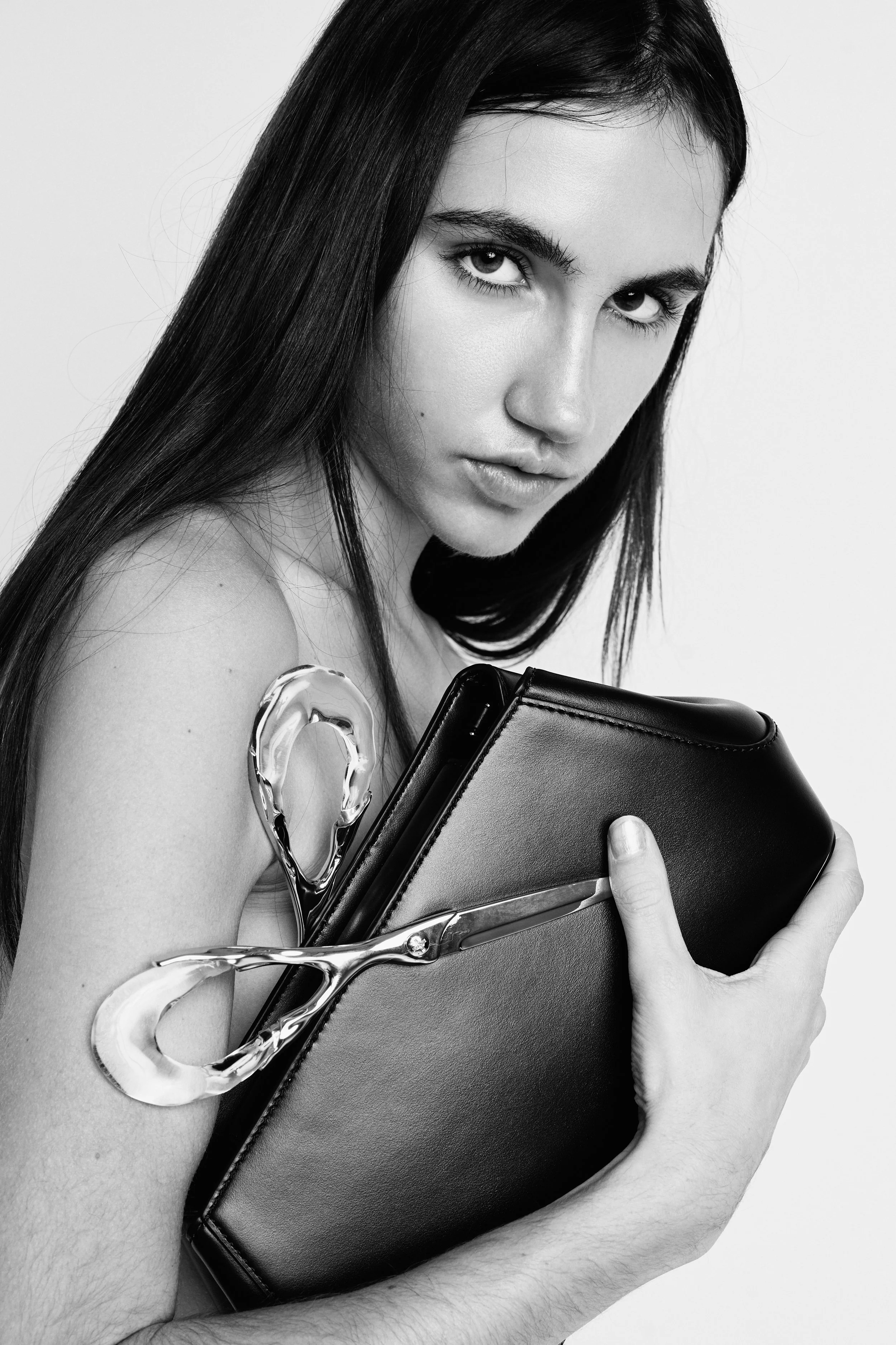 Black and white photo of a woman with long dark hair holding a black clutch purse, with a pair of large scissors near her shoulder and a piece of clothing hanging on her arm.