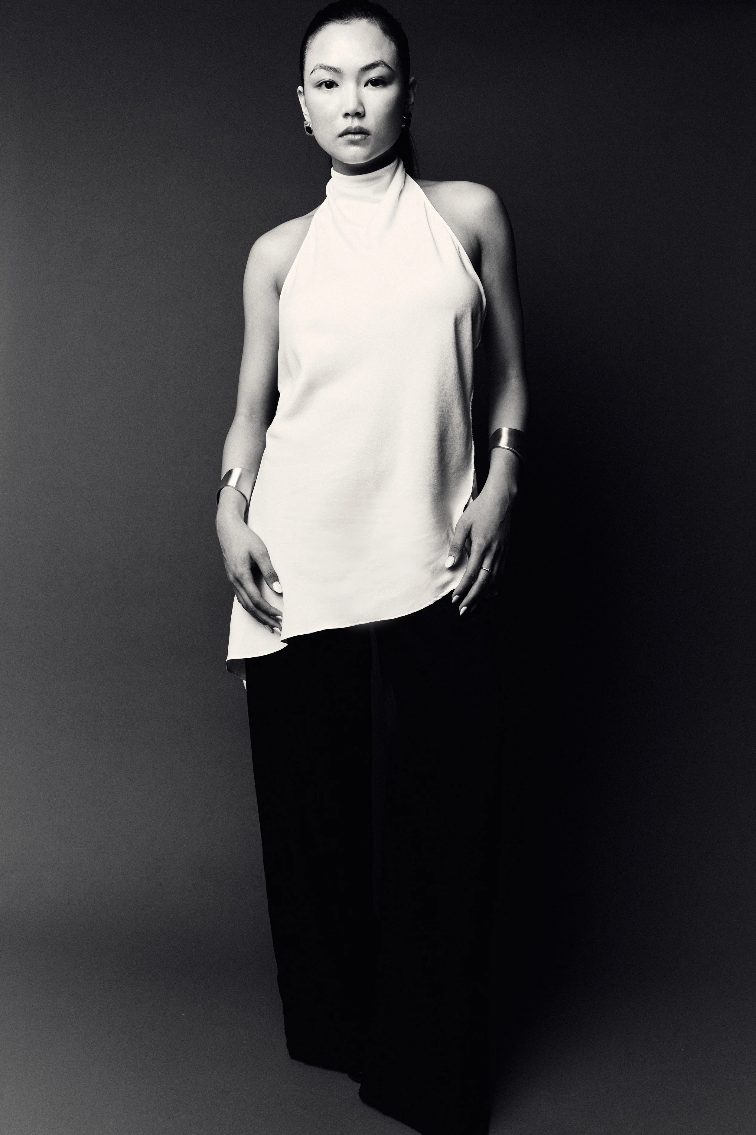 Black and white portrait of a woman with sleek hair, wearing a sleeveless high-neck top, accessorized with rings, earrings, a bracelet, and a choker, standing against a gradient background.