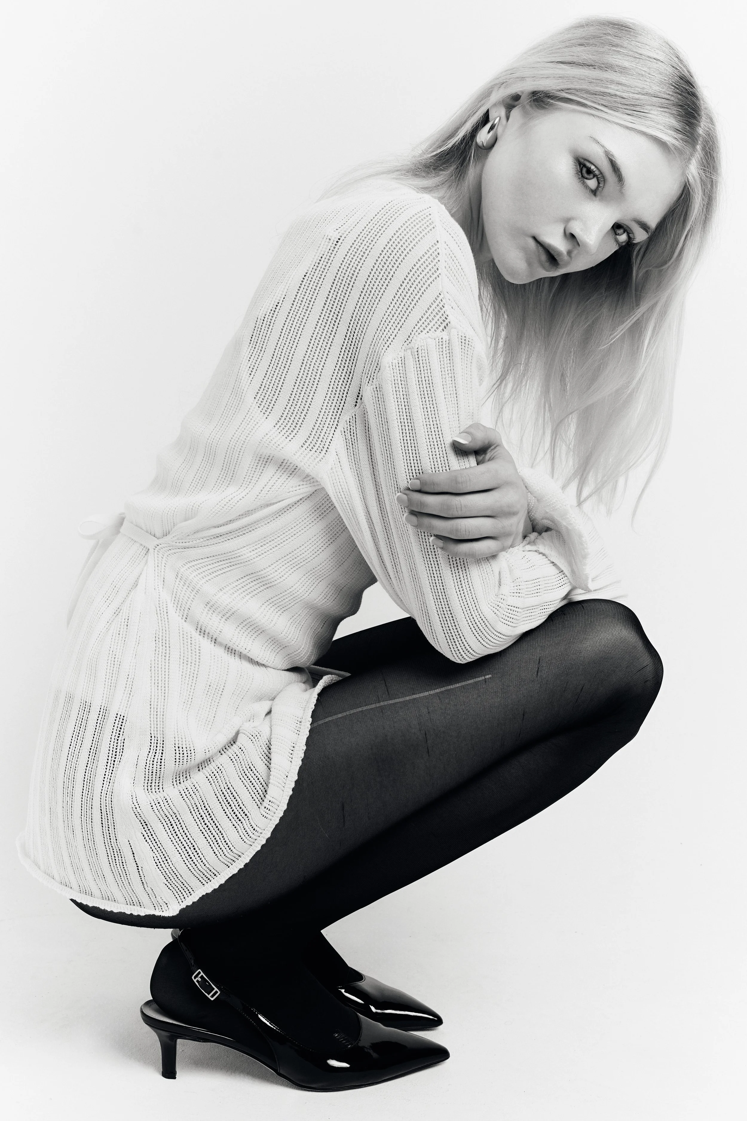 A woman with long blonde hair and light skin is wearing a light-colored, long-sleeve, knitted sweater and black tights with shiny black high-heeled shoes. She is sitting on her knees with her hands resting on her knees, looking directly at the camera with a slight smile, against a plain white background.