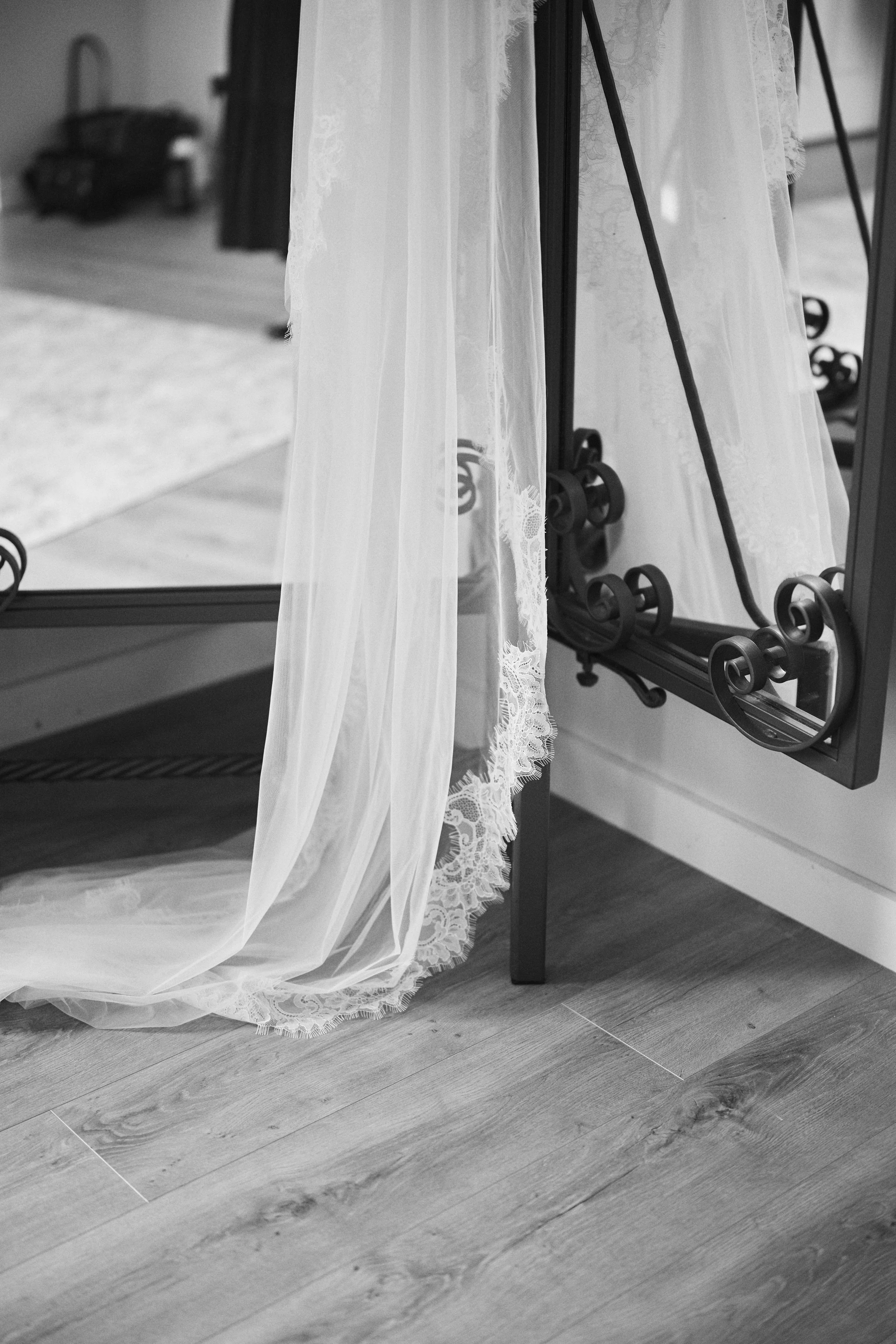 A close-up of a curtain and a mirror with a wrought iron frame, showing part of a room with a wooden floor and a blurred piece of furniture in the background.