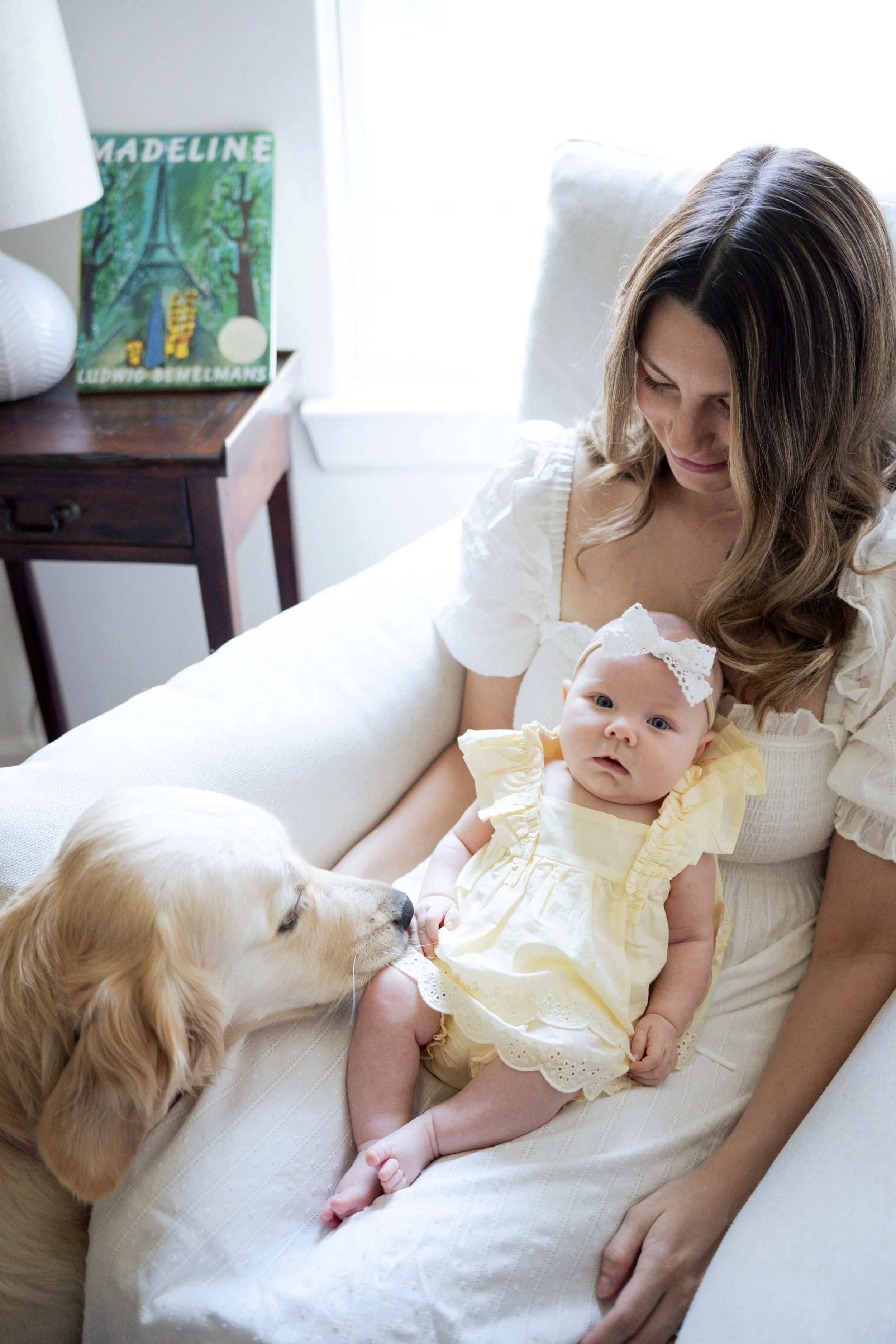 A woman holding a baby girl dressed in yellow, sitting on a white sofa with a golden retriever dog nearby. There is a nightstand with a lamp, a book titled 'Madeline' and a painting of the Eiffel Tower in the background.