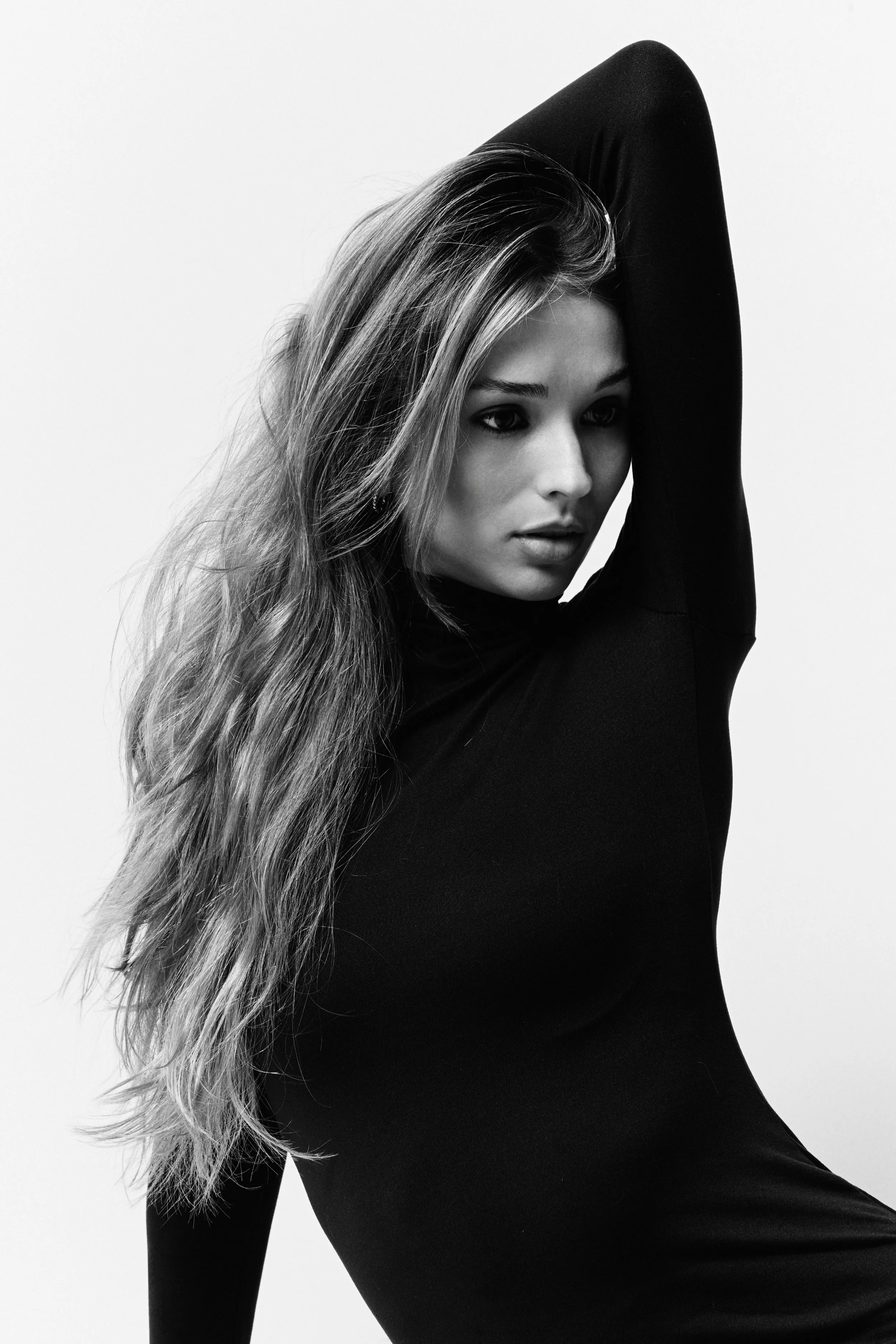 Black and white photo of a woman with long, wavy hair, wearing a black long-sleeve top, with one arm raised above her head, looking directly at the camera.