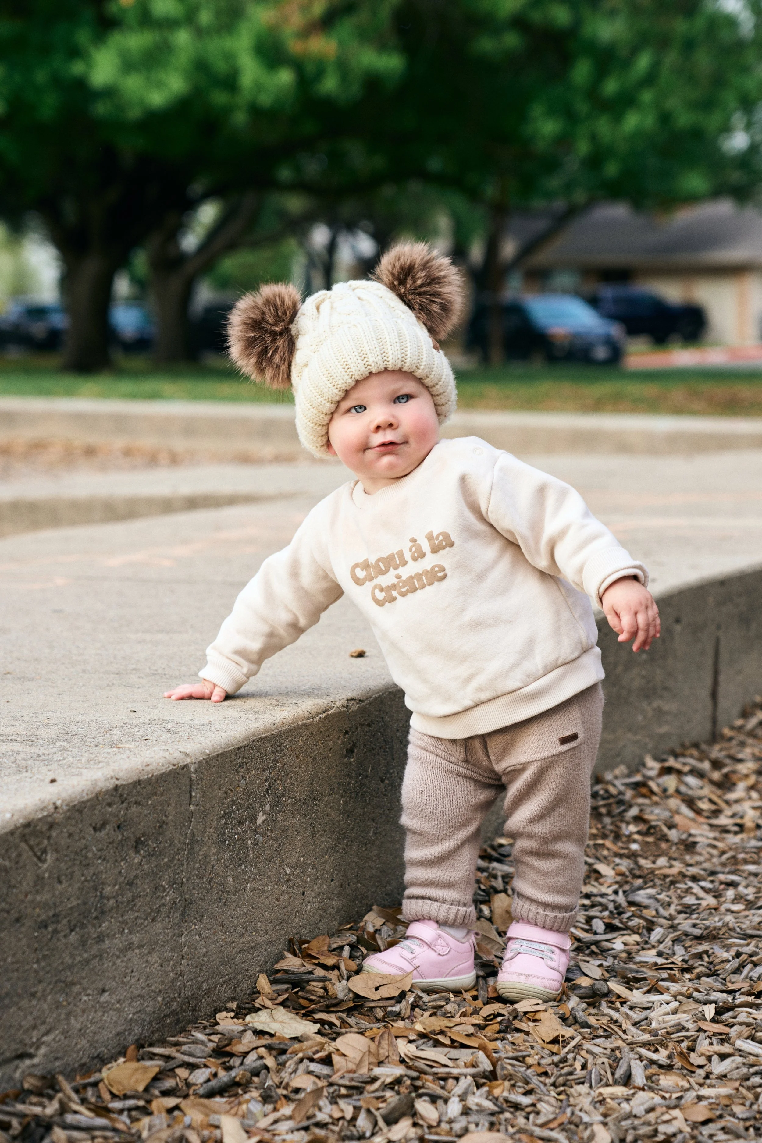 Young child outdoors wearing a beige knit hat with two pom-poms, a cream sweatshirt with French text, beige pants, and pink shoes, standing on a bed of leaves near a curb with trees and houses in the background.