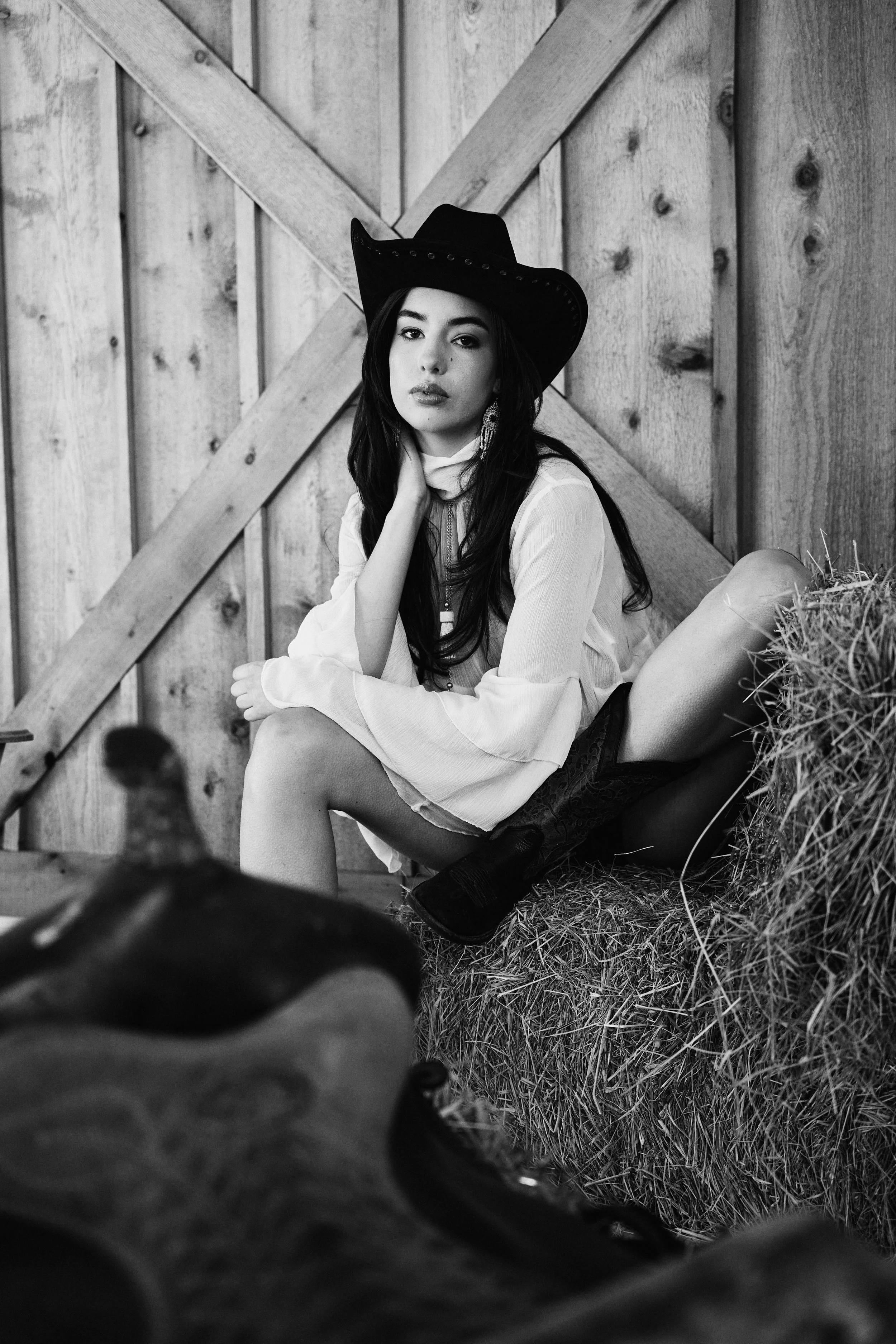 A woman is sitting against a wooden wall in a barn-like setting, wearing a cowboy hat and earrings, with her hand resting near her face, and looking at the camera. The photo is in black and white.