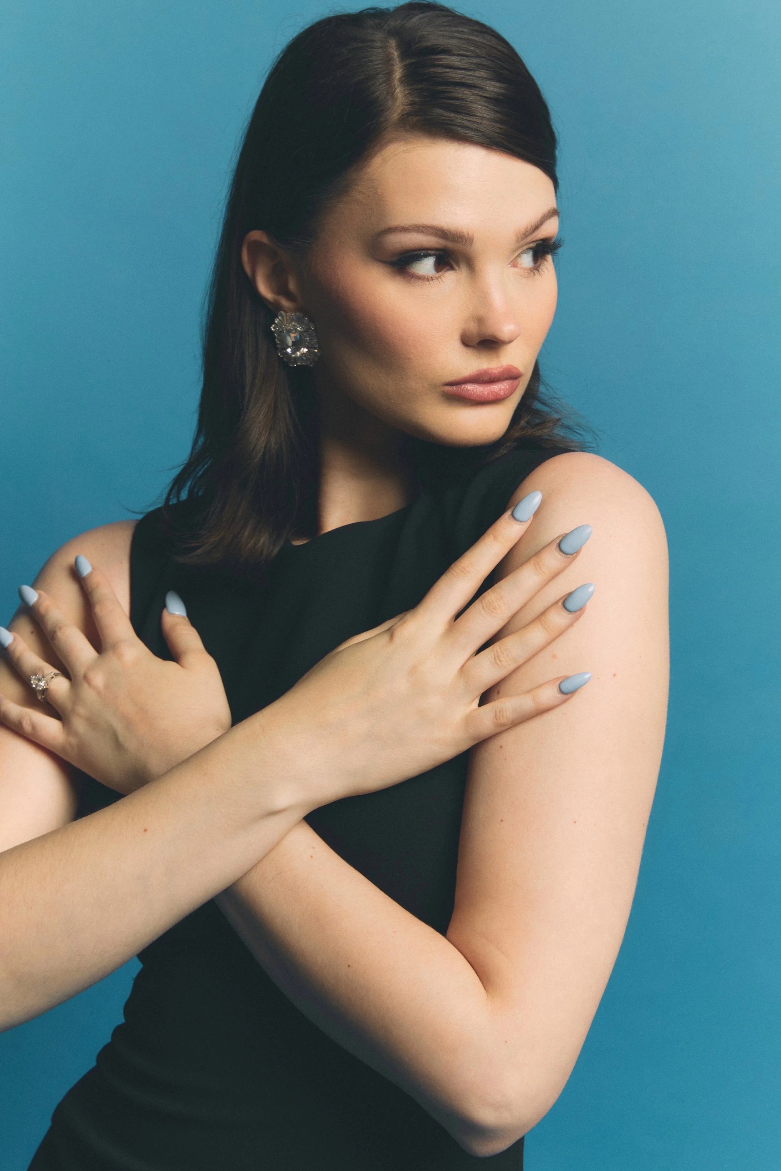 A woman with dark brown hair, wearing a black sleeveless top, large decorative earrings, and light blue nail polish, looking to the side against a solid blue background, with her arms crossed over her chest.