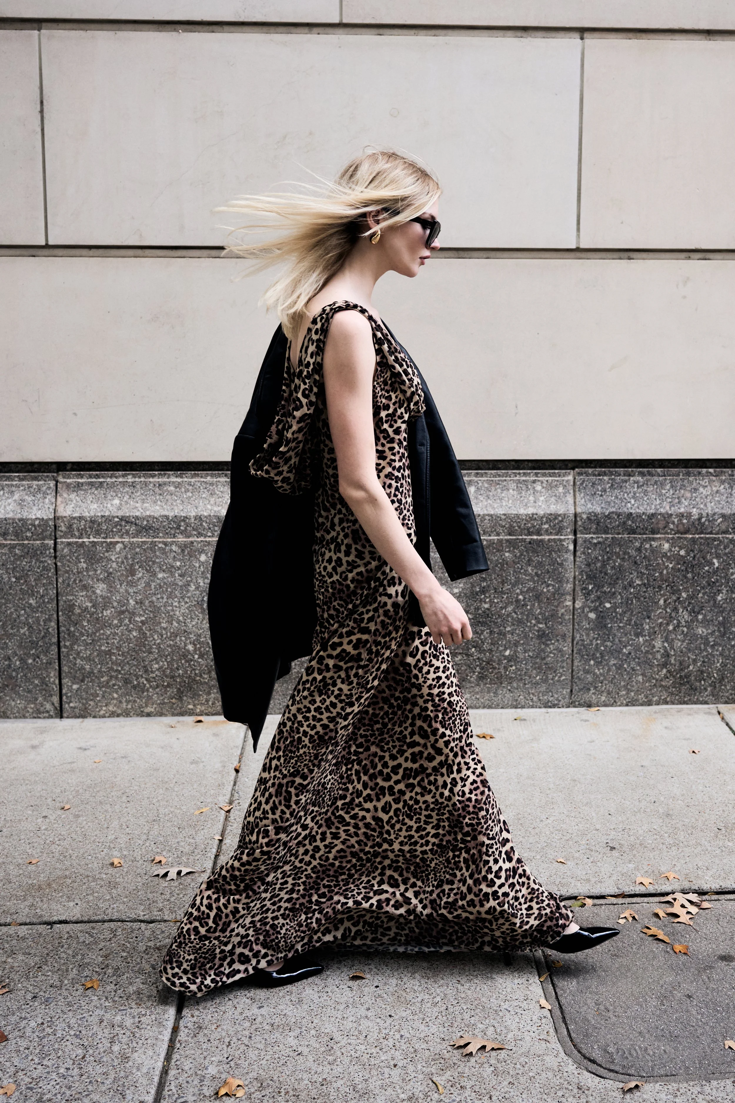 A woman walking on the sidewalk wearing a long leopard print dress, black shoes, sunglasses, with a black jacket draped over her shoulder, and her blonde hair blowing in the wind.