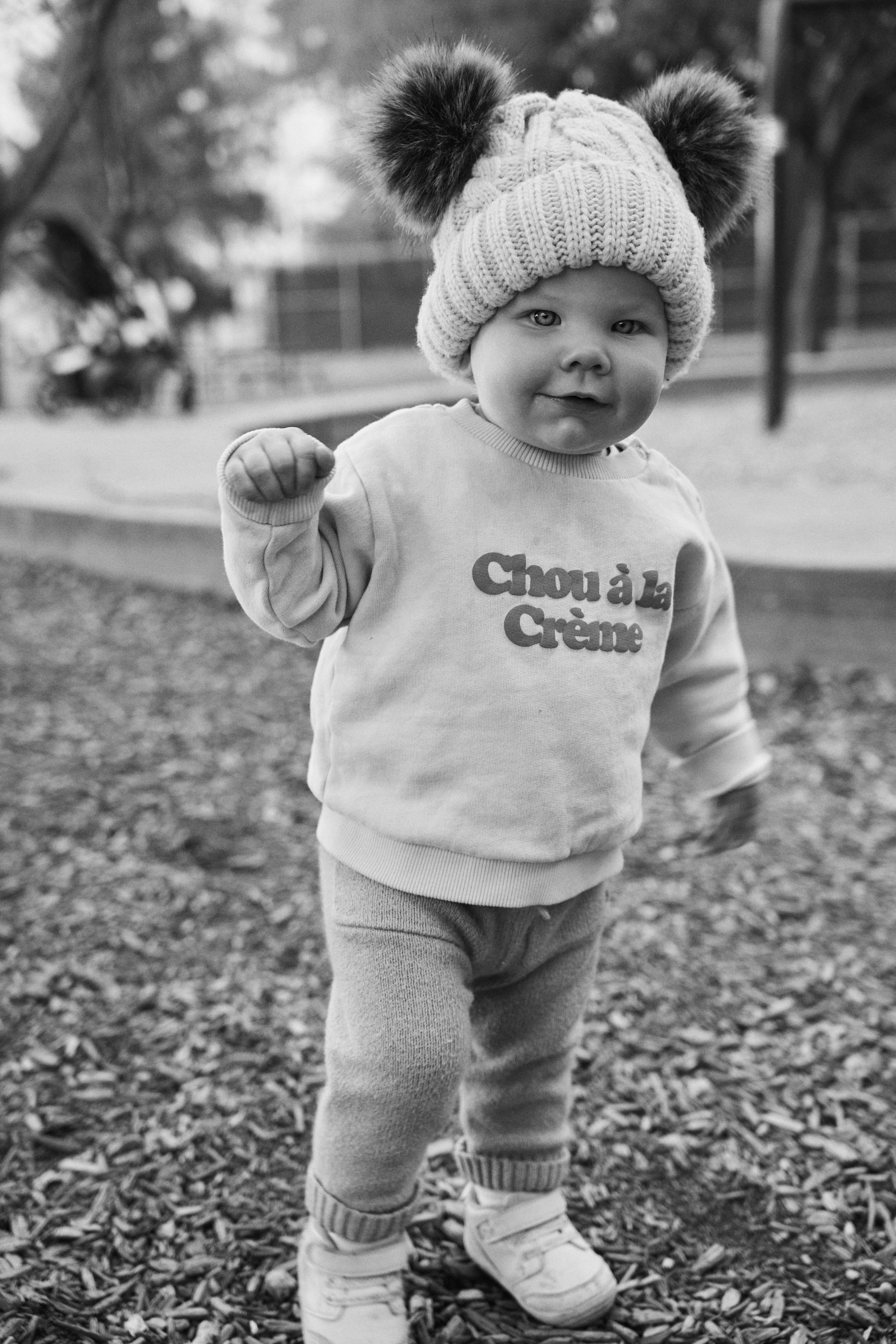 Young child outdoors wearing a knit hat with pom-poms, sweatshirt, pants, and sneakers. The child is holding one fist up and smiling, with a background of trees and leaves.