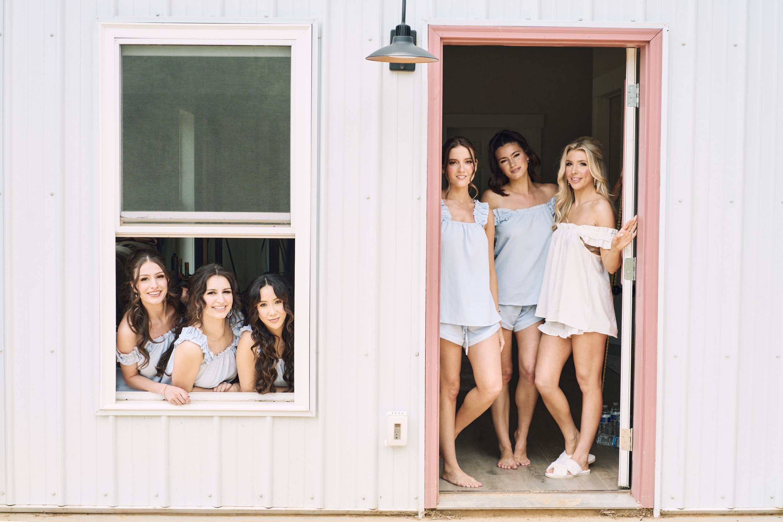 A group of women in matching light blue and white sleepwear standing in an open doorway and a window of a white building, smiling at the camera.