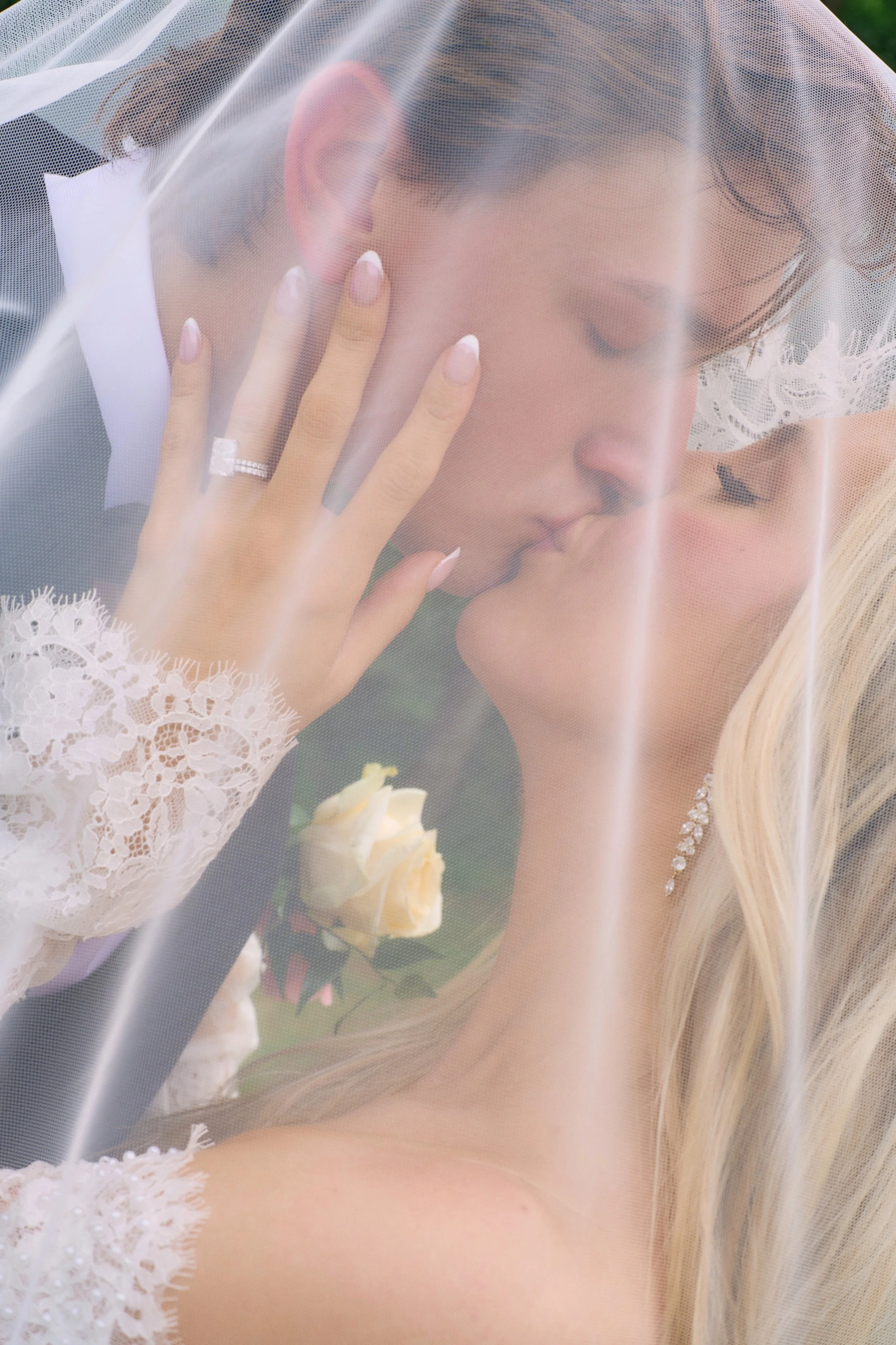 A bride with long blonde hair, wearing a white dress with lace details, is behind a sheer veil. She is holding her hand to her face, showing a diamond ring on her finger, and appears to be in a contemplative or emotional moment.