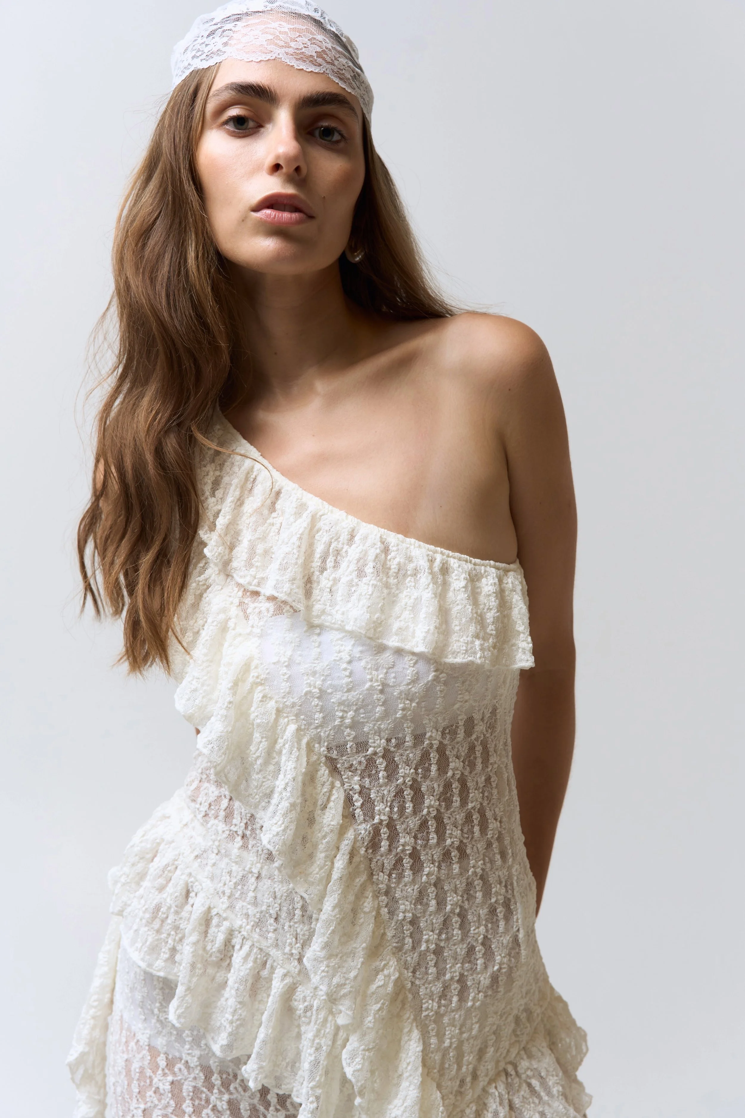 A woman with long wavy brown hair wearing a white lace off-shoulder dress and a white lace headband against a plain background.