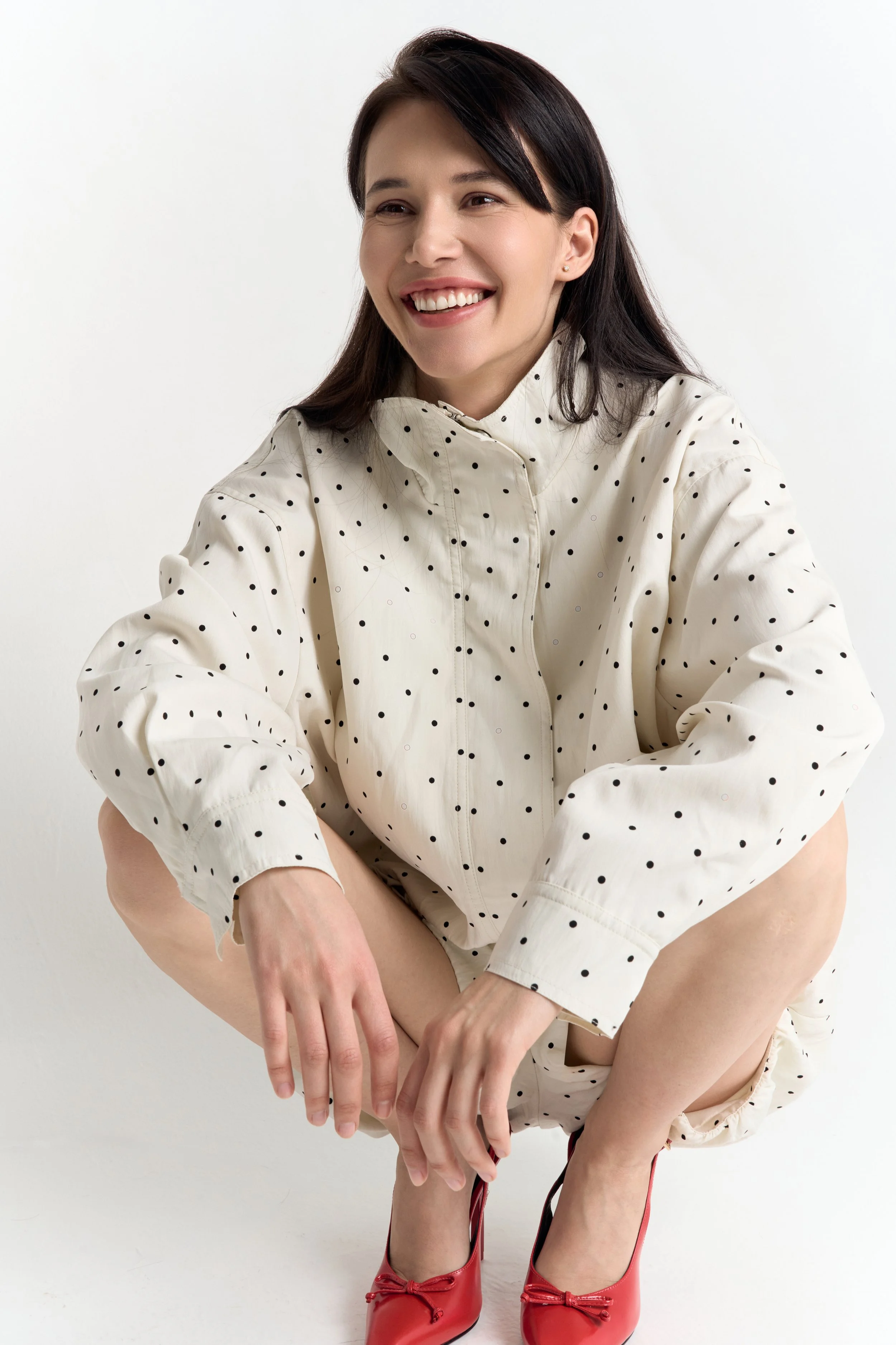 A woman with dark brown hair and fair skin, smiling with teeth visible, is sitting with her arms resting on her knees. She is wearing a cream-colored shirt with black polka dots and red high heels. The background is plain white.