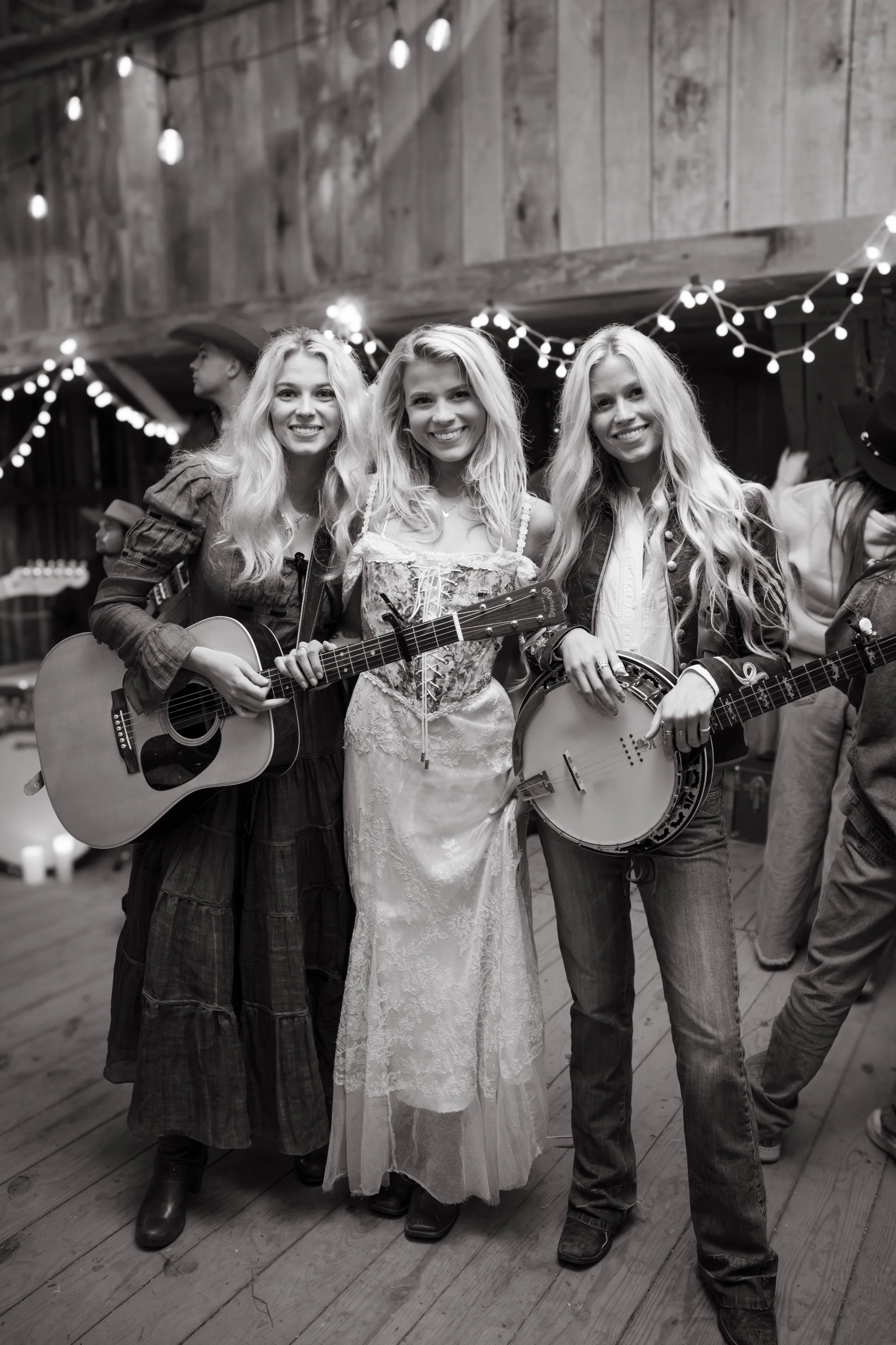 Three women in a barn with string lights, holding guitars and smiling at the camera.