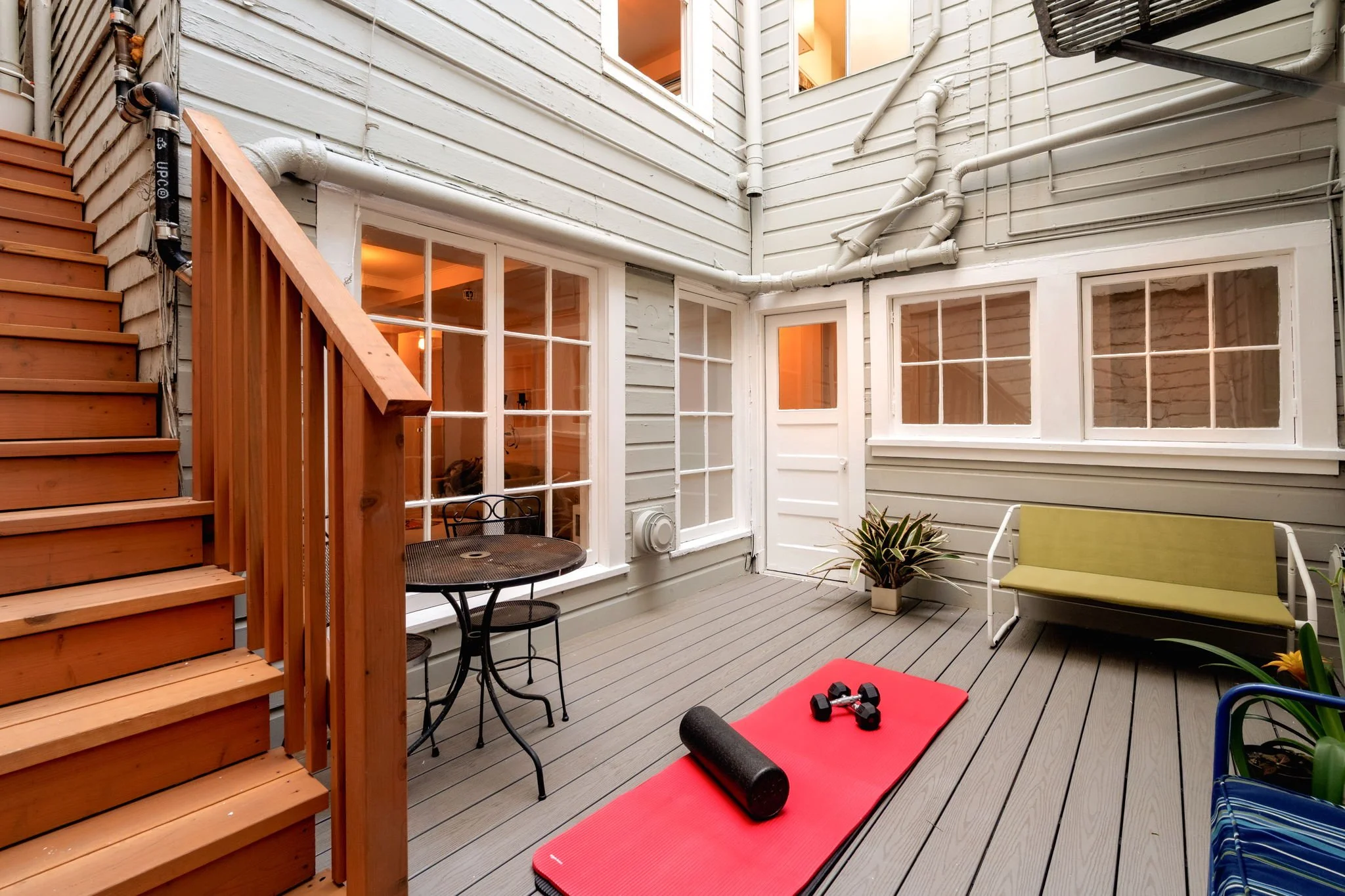 A cozy outdoor patio or balcony area with a red exercise mat, dumbbells, a foam roller, and a black foam roller on the floor, surrounded by furniture and potted plants, with house windows and a staircase leading up to the space.