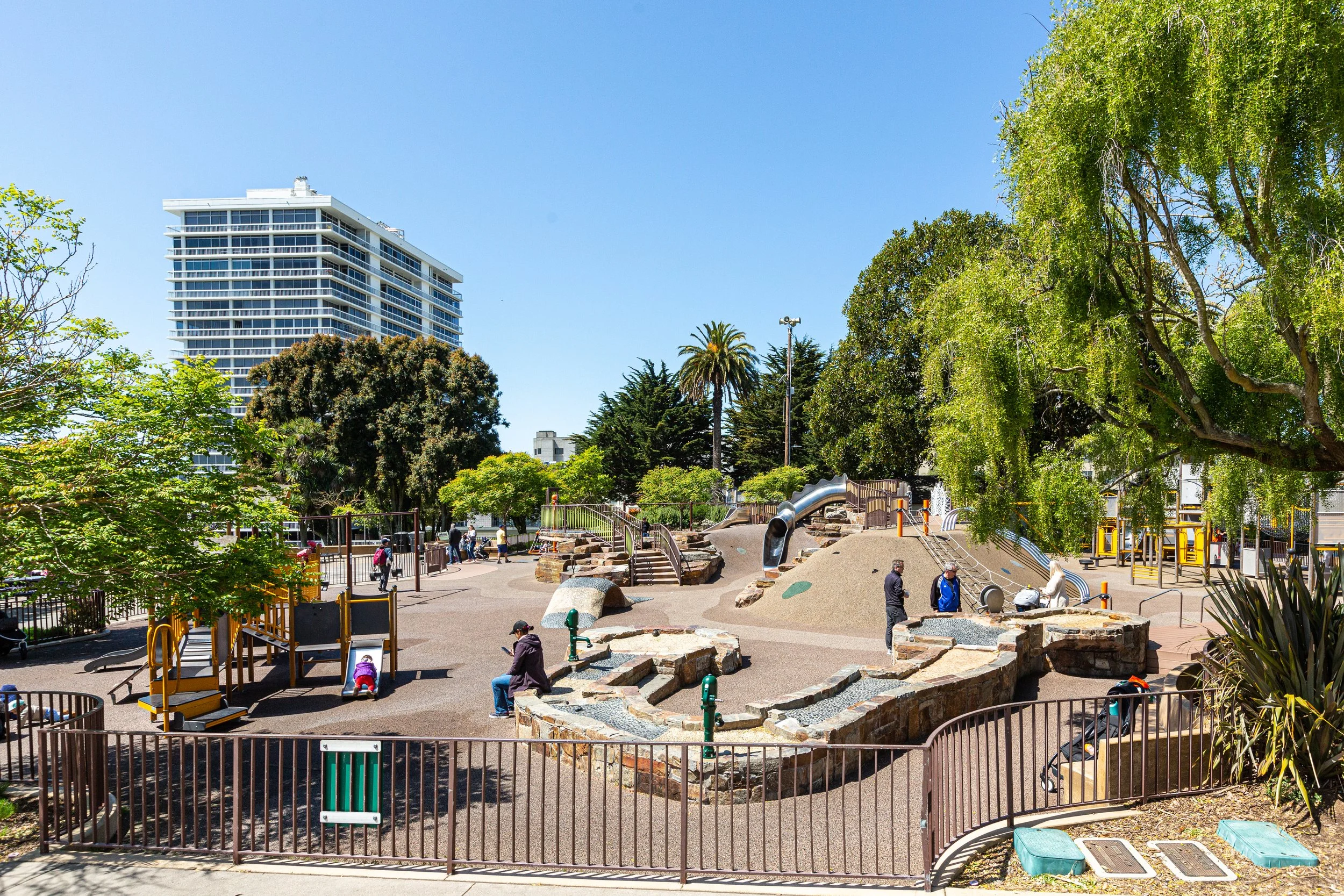 Public park with playground equipment, trees, and a tall modern building in the background under a clear blue sky.