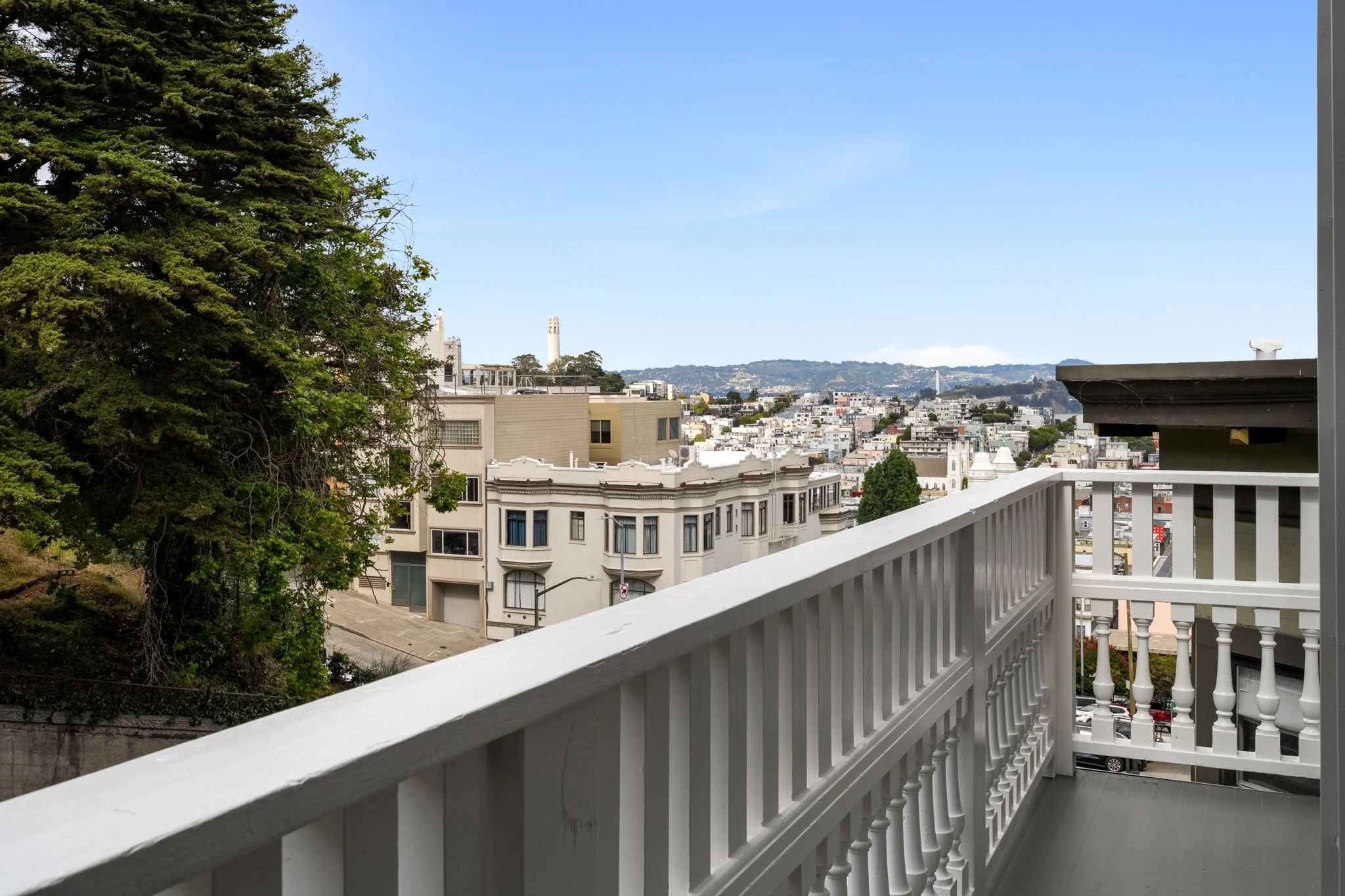 Urban cityscape view from a balcony with a white railing, showing buildings, trees, and a hilly background under a clear blue sky.