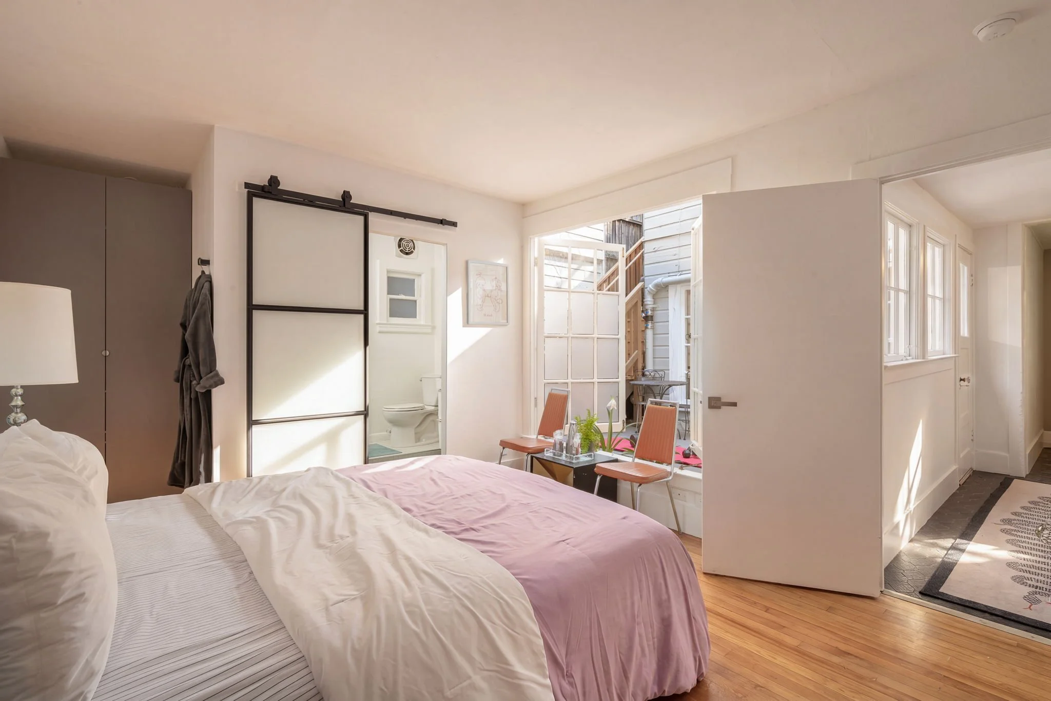 A bedroom with a bed, bedside lamp, and a view into a bathroom and small outdoor area through an open door. The room has light-colored walls and wooden flooring.