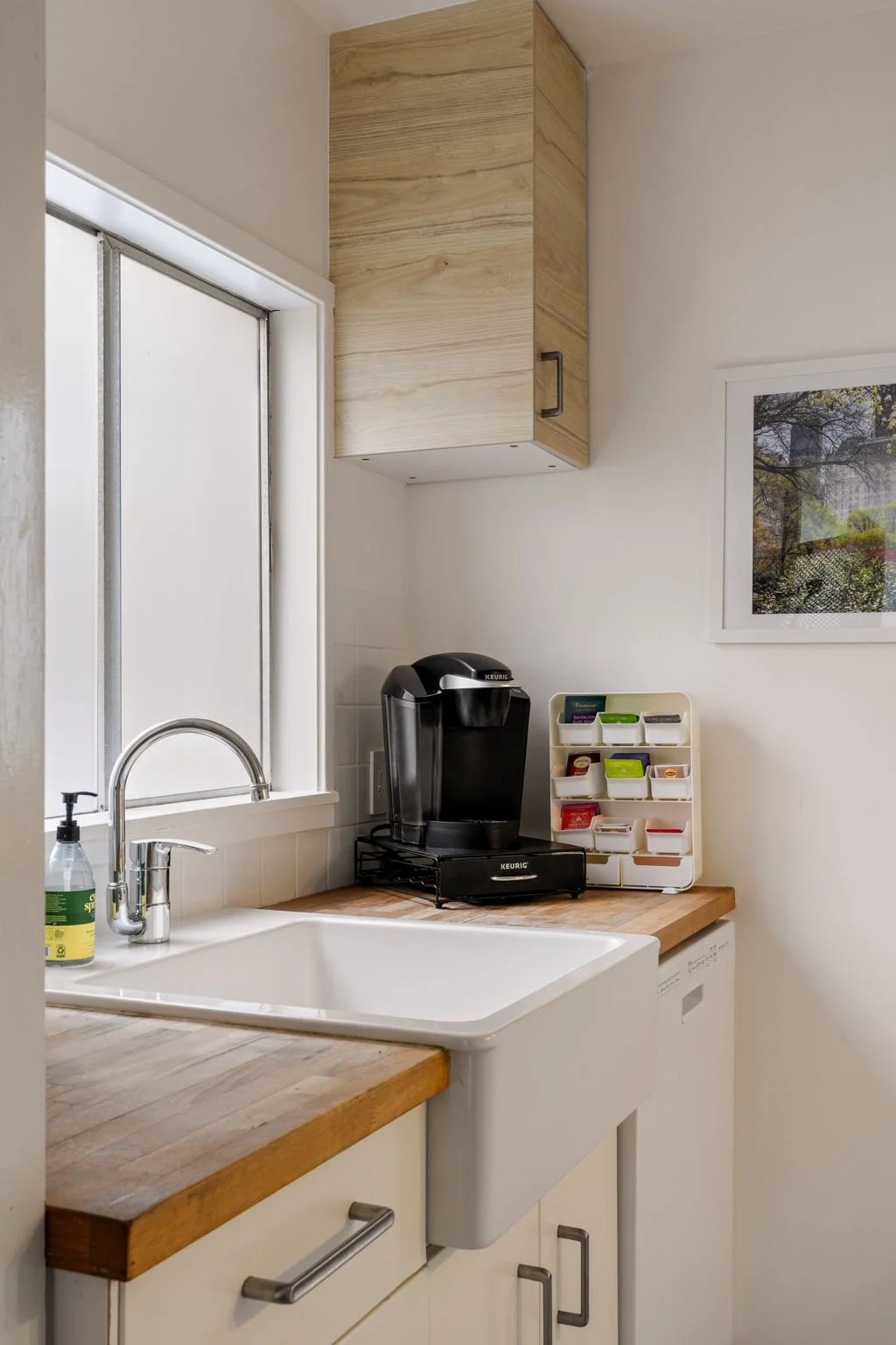 Kitchen with a white sink, wooden countertop, Keurig coffee maker, spice rack, window, wall cabinet, framed picture, and dishwasher.