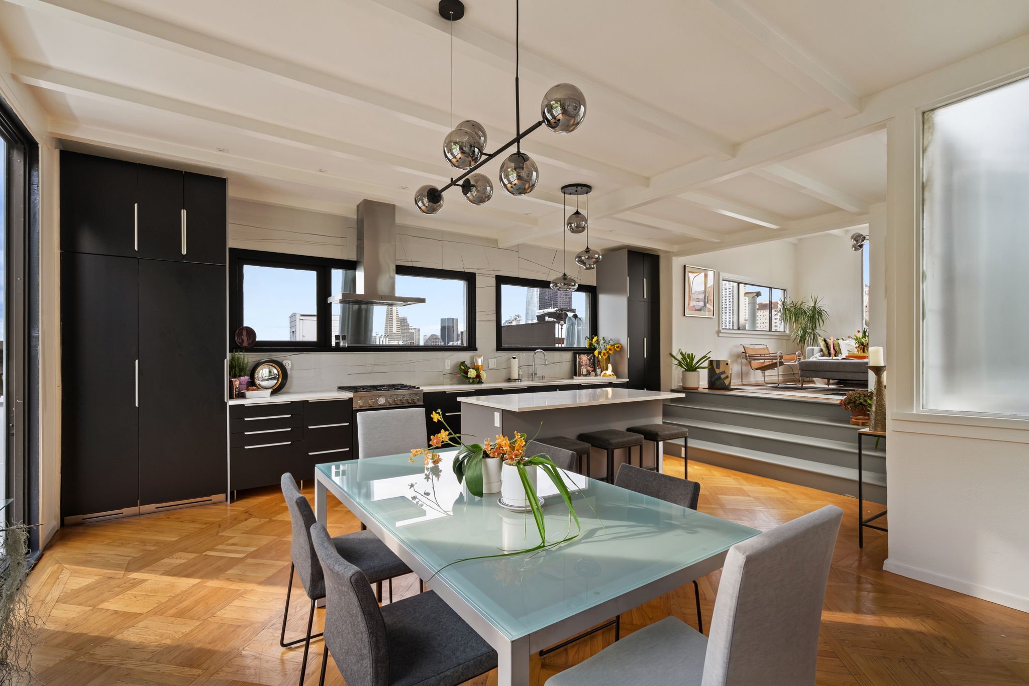 Modern open-concept kitchen and living room with black cabinetry, white countertop, dining table with six chairs, and large windows showing city skyline.