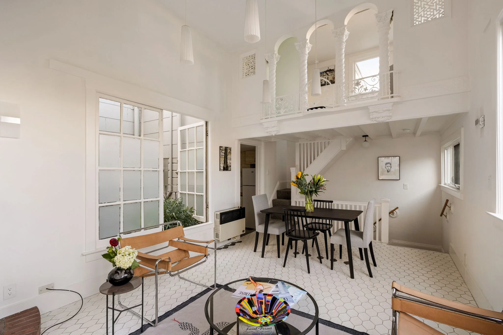 Interior view of a white living room with a high ceiling, tall windows, a dining table with chairs, a small table with flowers, and various decorative elements.